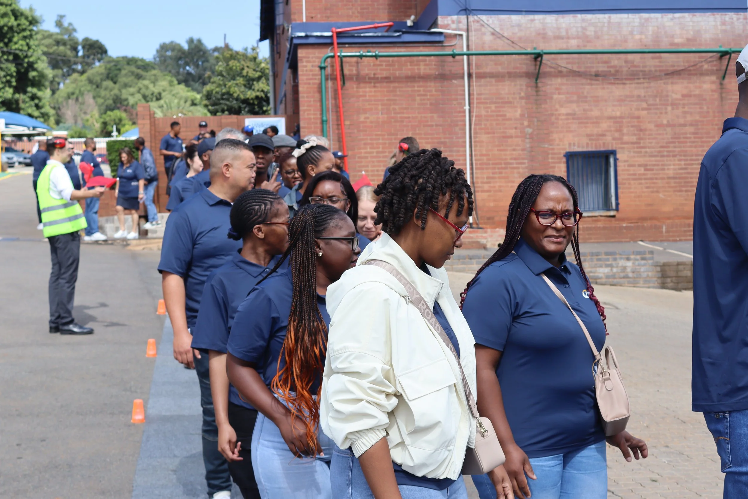 A line of people standing outdoors on a sidewalk during a daytime event, with some wearing blue shirts and headphones, and a few in casual clothing, near traffic cones, with a brick building and green trees in the background.