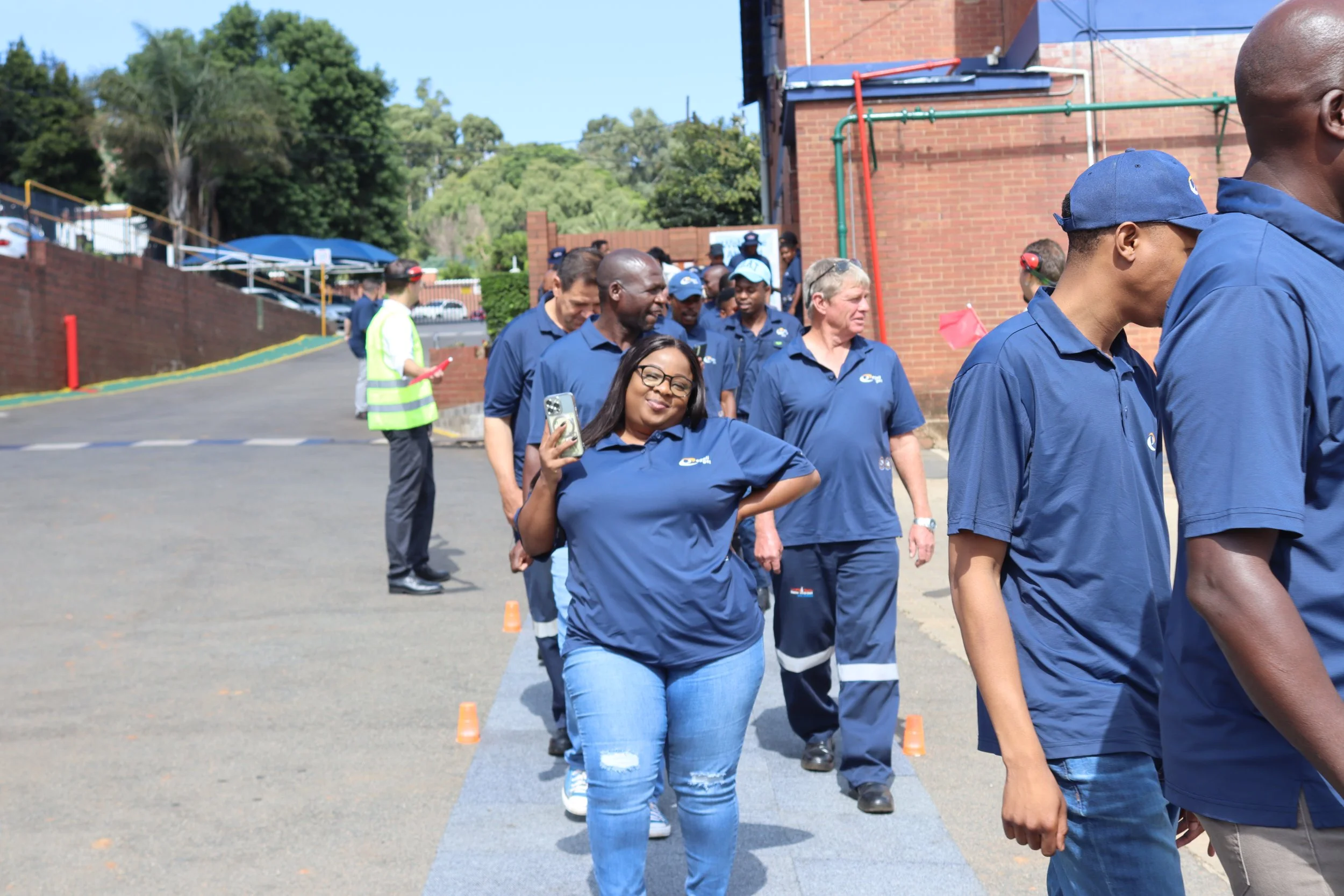 Group of diverse people dressed in blue shirts participating in an outdoor team-building activity involving walking in a line between small orange cones.