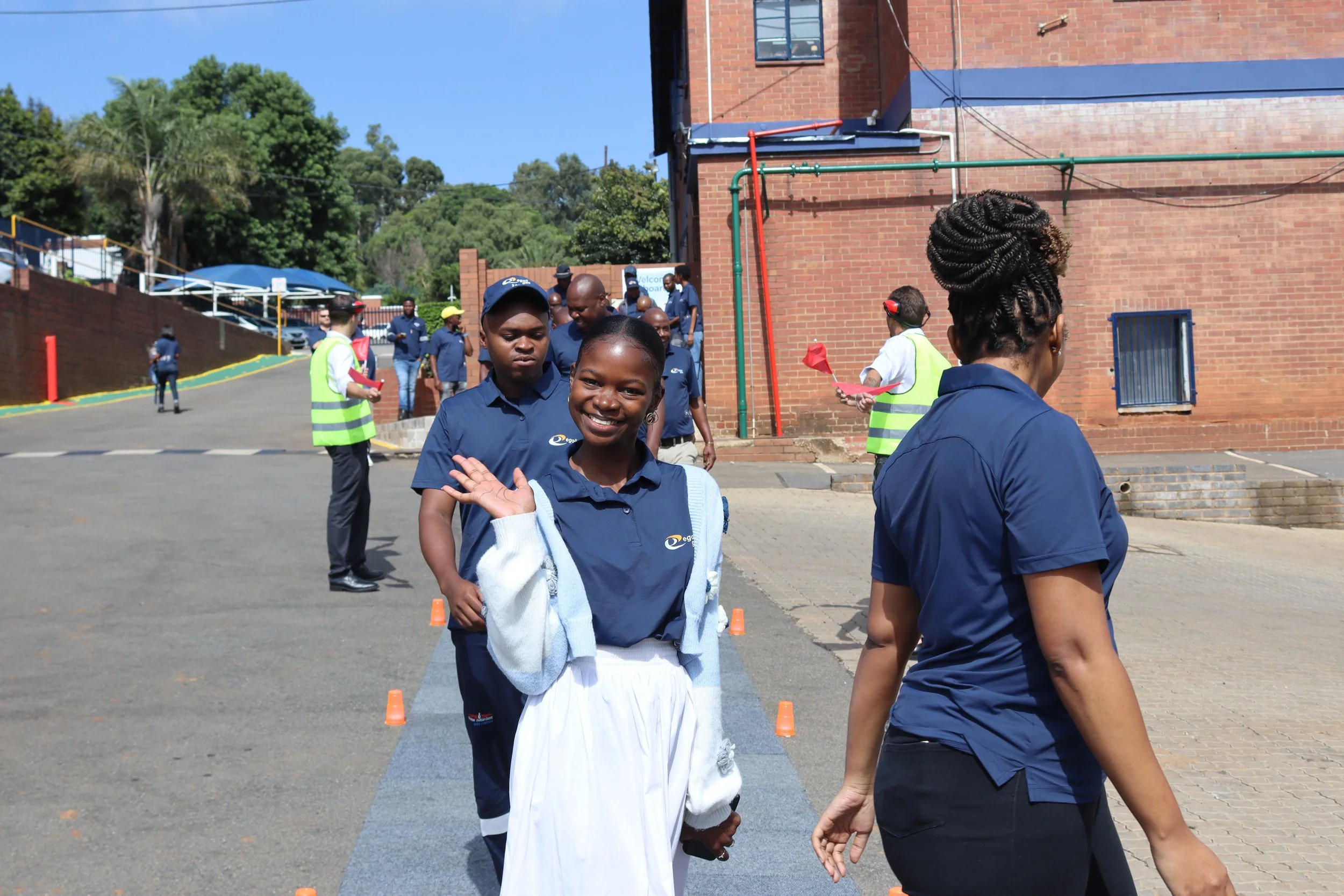 Group of people, mainly young women, standing outdoors near a brick building on a sunny day. Some are dressed in blue uniforms, and there are orange cones on the ground, indicating some form of organized activity or event.