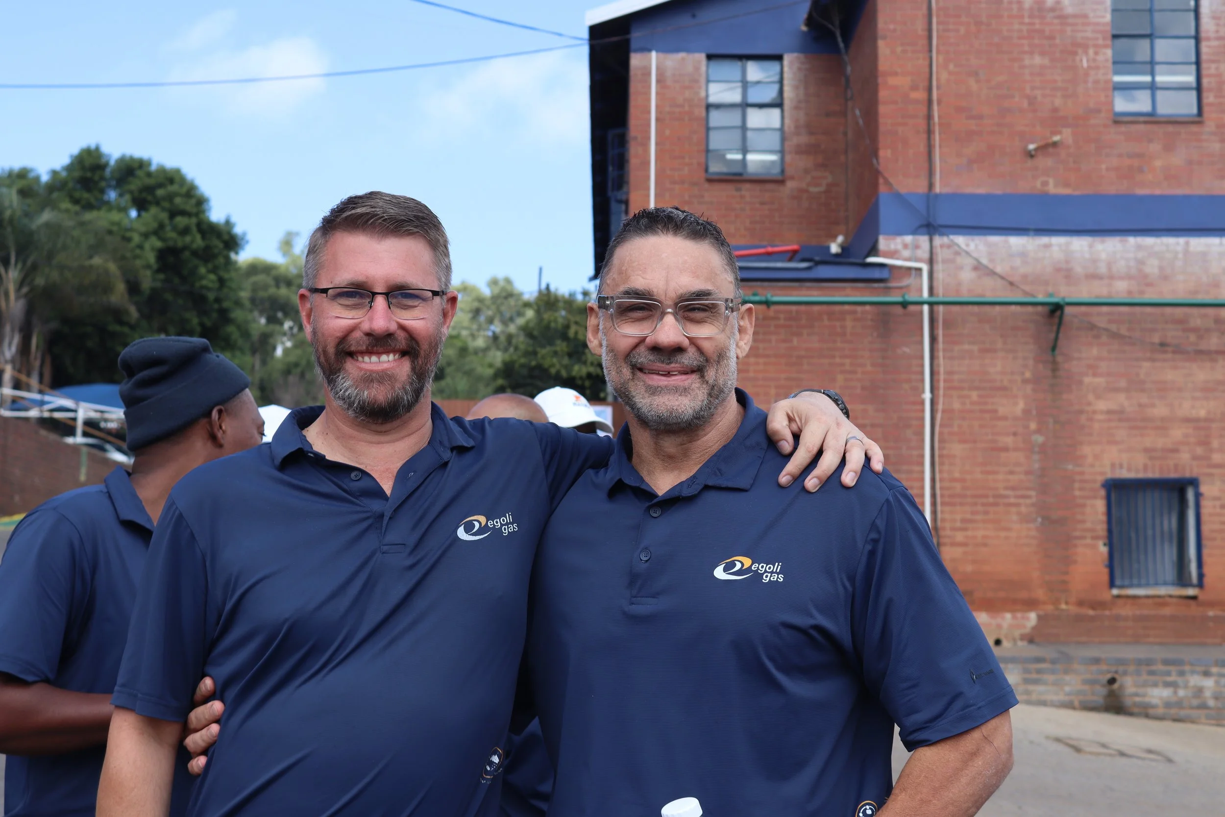 Two men smiling and standing close together, wearing matching blue shirts with a company logo, outdoors in front of a brick building.