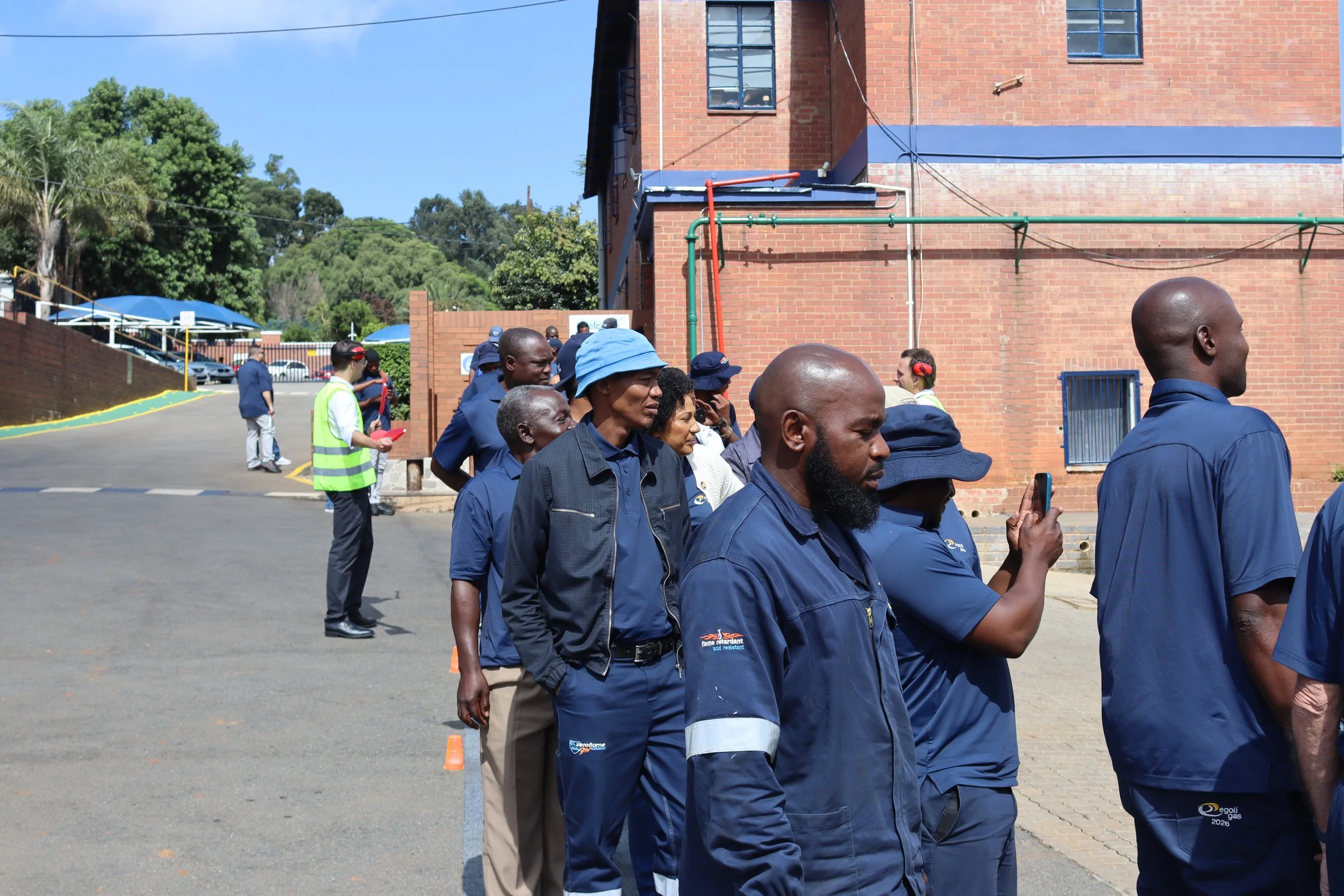 People lining up outdoors in front of a brick building, some wearing uniforms and hats, with trees and a parking lot in the background.