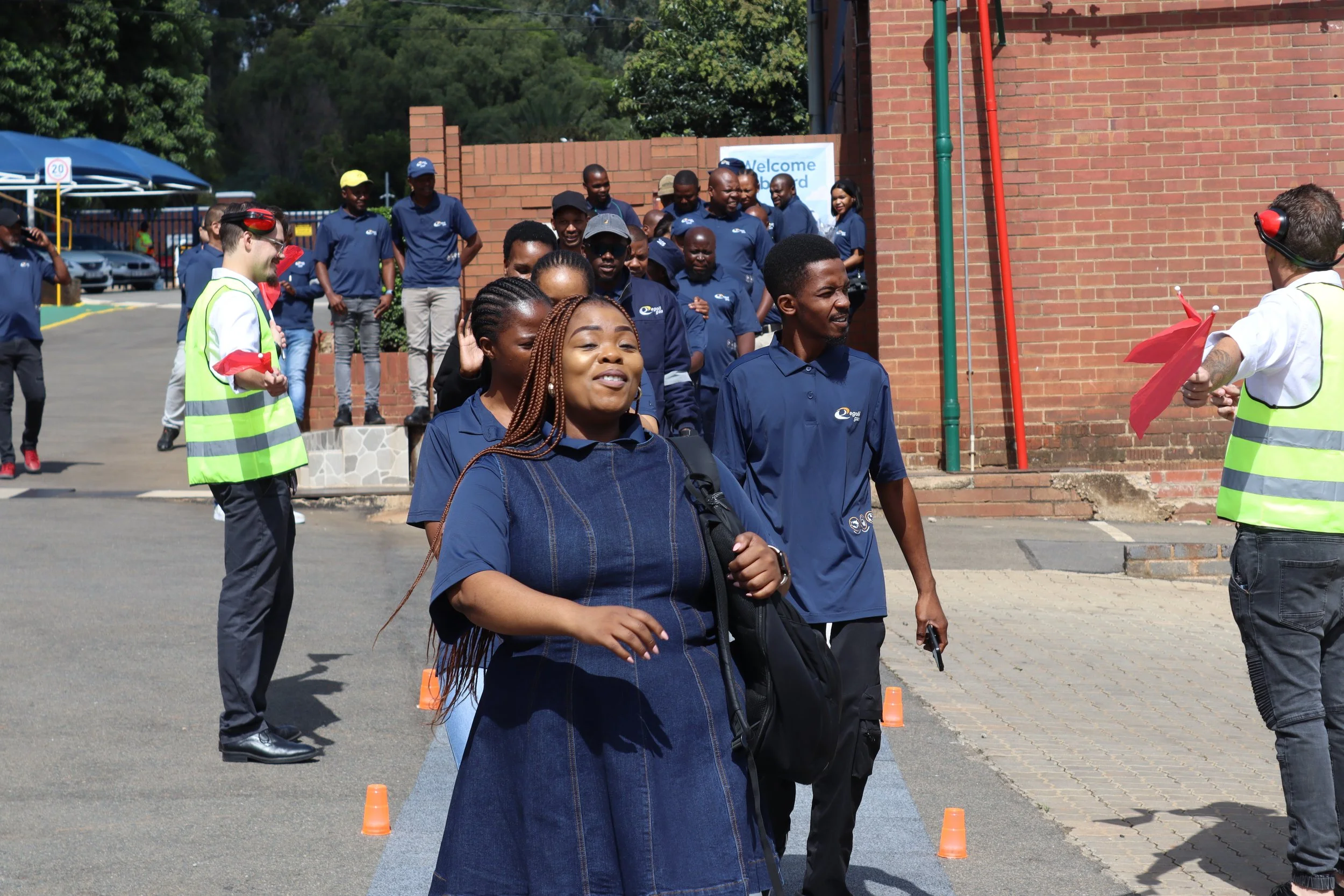 Group of people walking outdoors near brick building, some wearing blue uniforms, one person wearing a yellow safety vest, and some wearing headphones, on a sunny day.