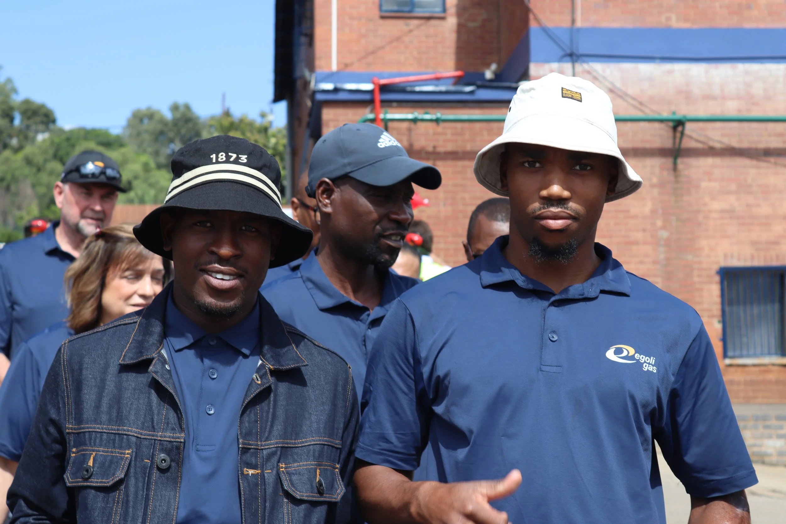 Group of people outdoors, including two men wearing navy blue shirts and hats, walking and talking in front of a brick building with a blue sky background.