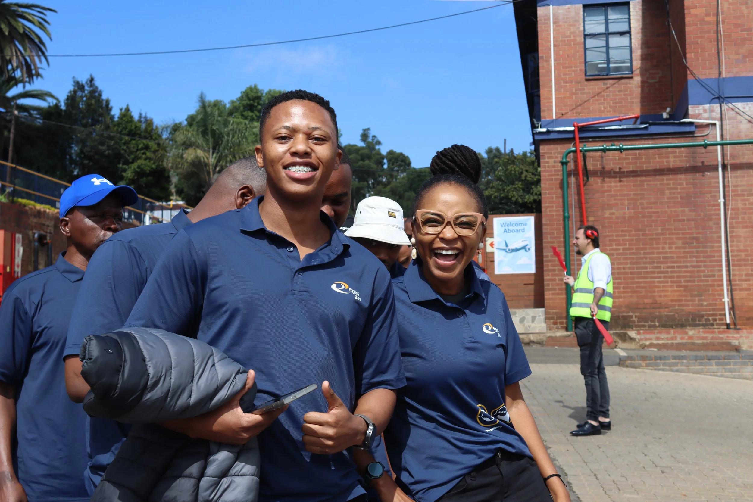 Group of smiling people wearing blue shirts outdoors with trees and buildings in the background.