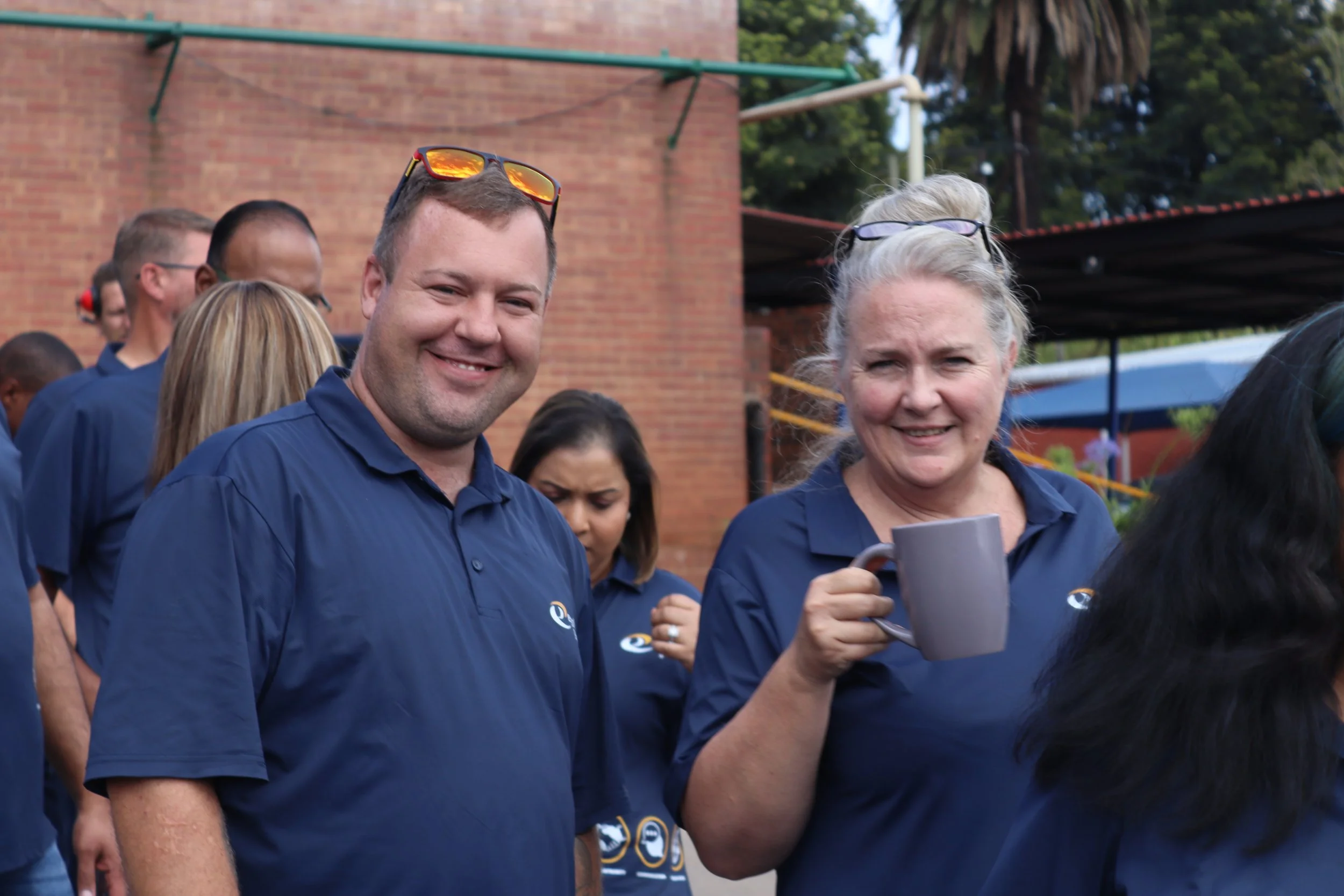 Group of people outdoors, two smiling adults in the foreground, wearing navy blue shirts, with others in the background, in front of a brick building and trees.