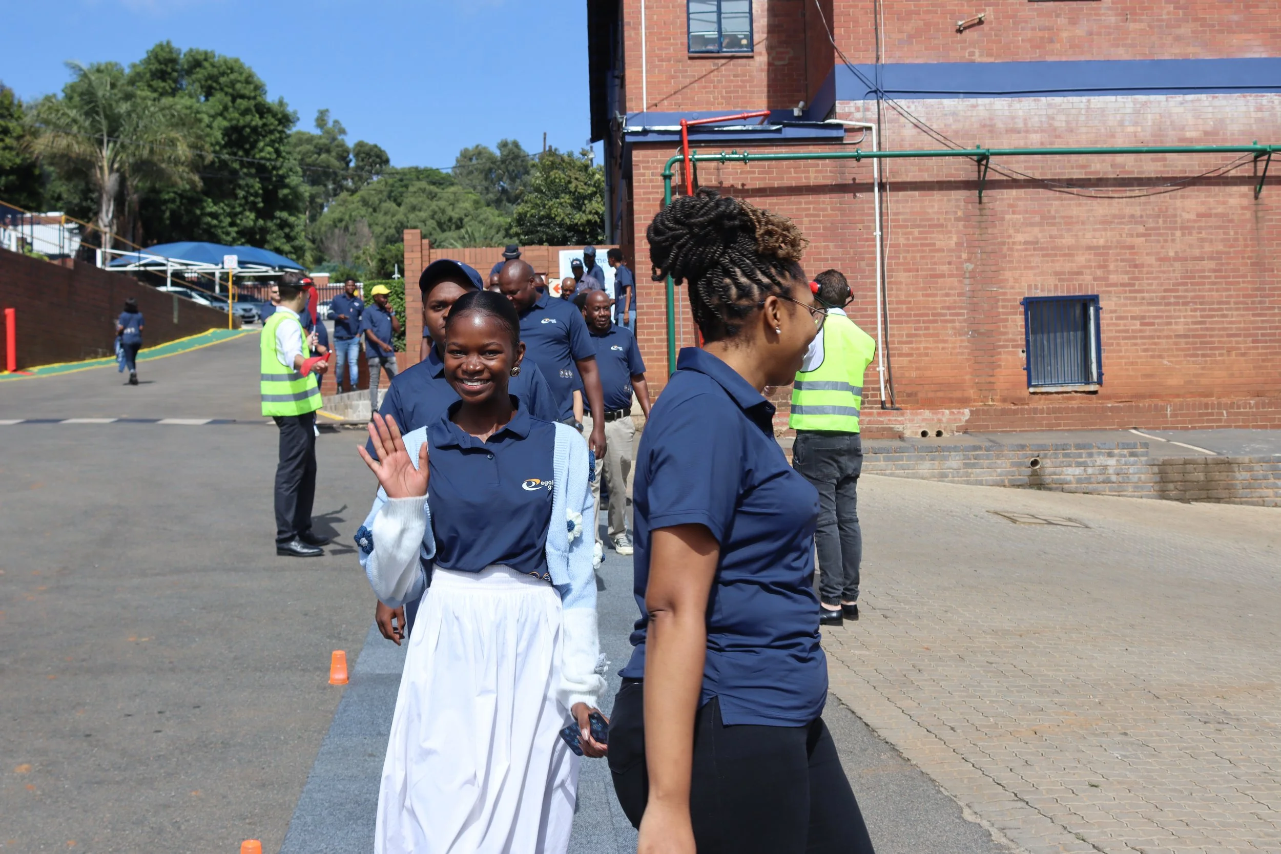 People gathered outdoors near a brick building, some smiling and waving at the camera, while others are engaged in an activity or conversation, with trees and a blue sky in the background.