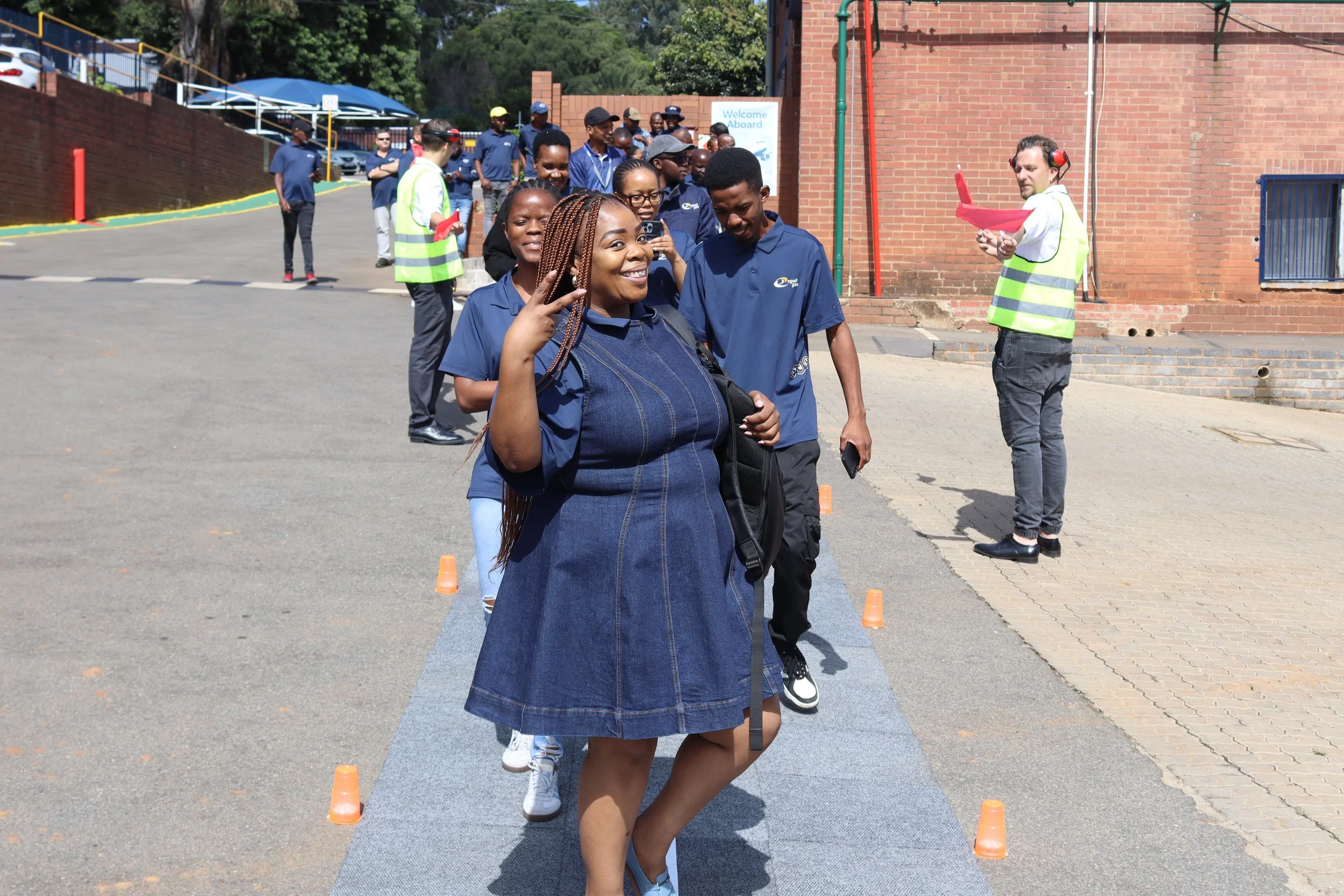 A group of people lined up outdoors, participating in an activity, with women and men, some wearing blue uniforms, and a woman at the front smiling and making a peace sign. There are orange cones and a ramp on the ground, with brick walls and a sign 