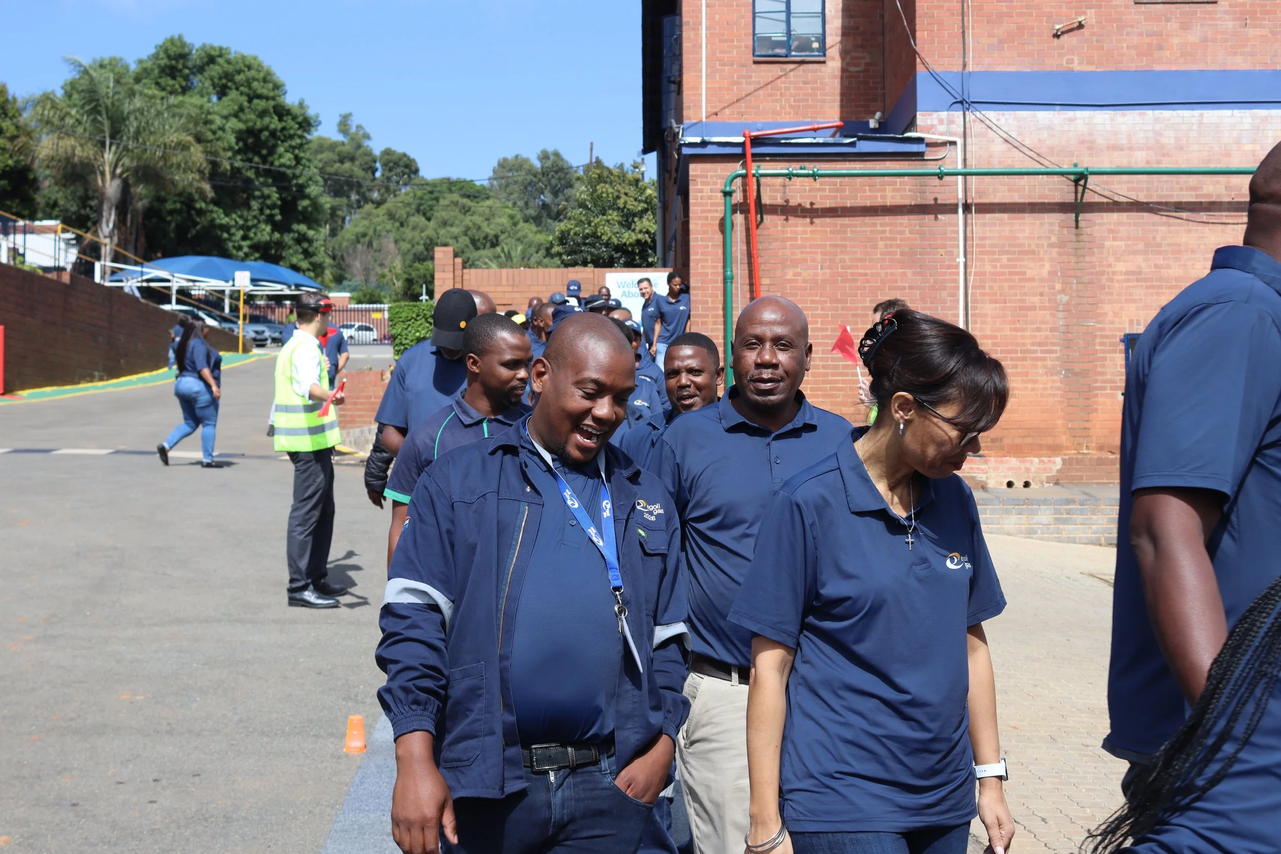 A group of diverse people dressed in blue uniforms walking in a line outdoors on a sunny day, with trees and a brick building in the background.