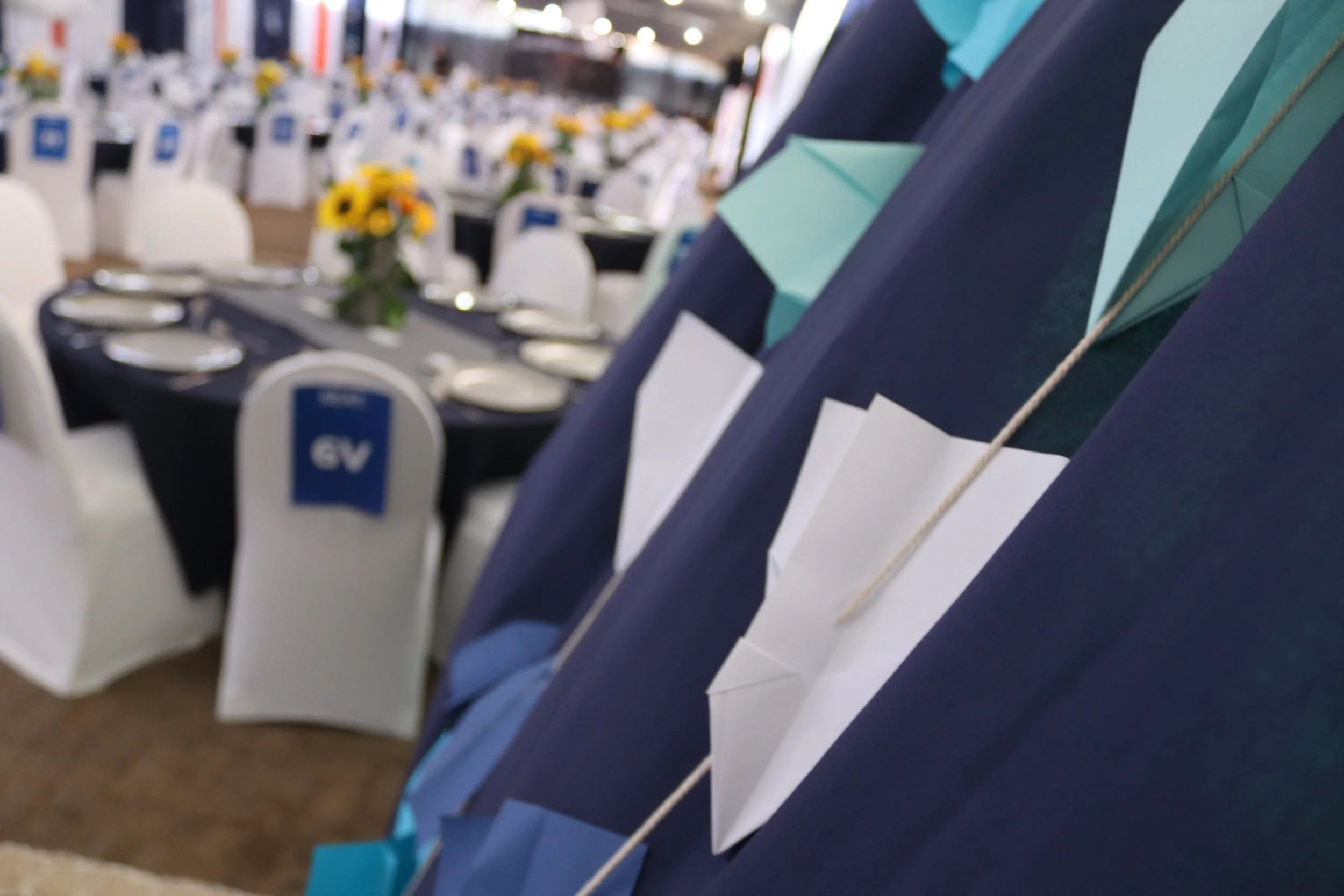 Close-up of folded paper napkins in blue and teal colors with a white tag, part of a display in a dining or event setting; blurred background showing arranged tables with white chairs, table settings, and floral centerpieces.