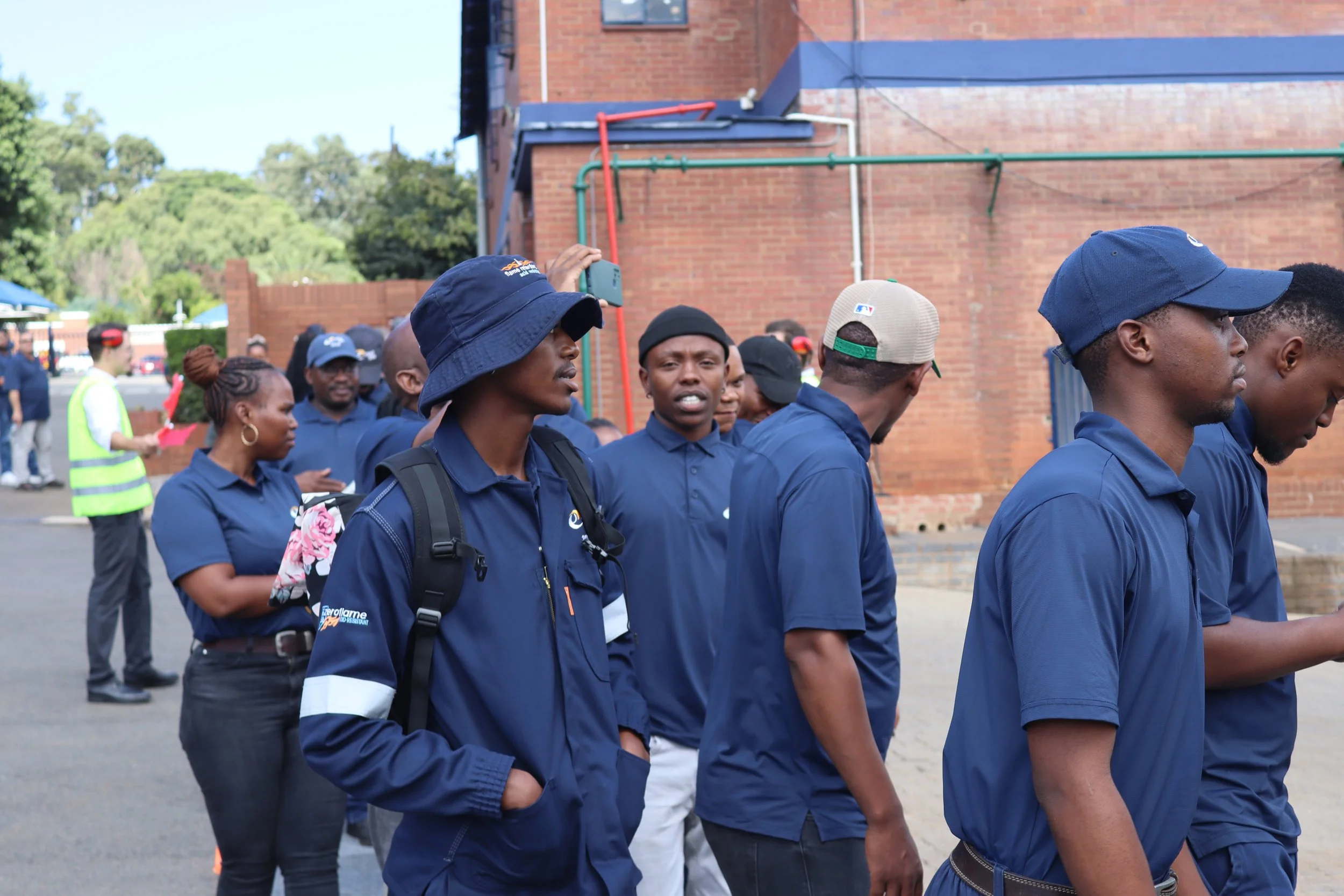 Group of people walking outdoors near a brick building, some wearing blue hats and jackets, with trees and a blue sky in the background.