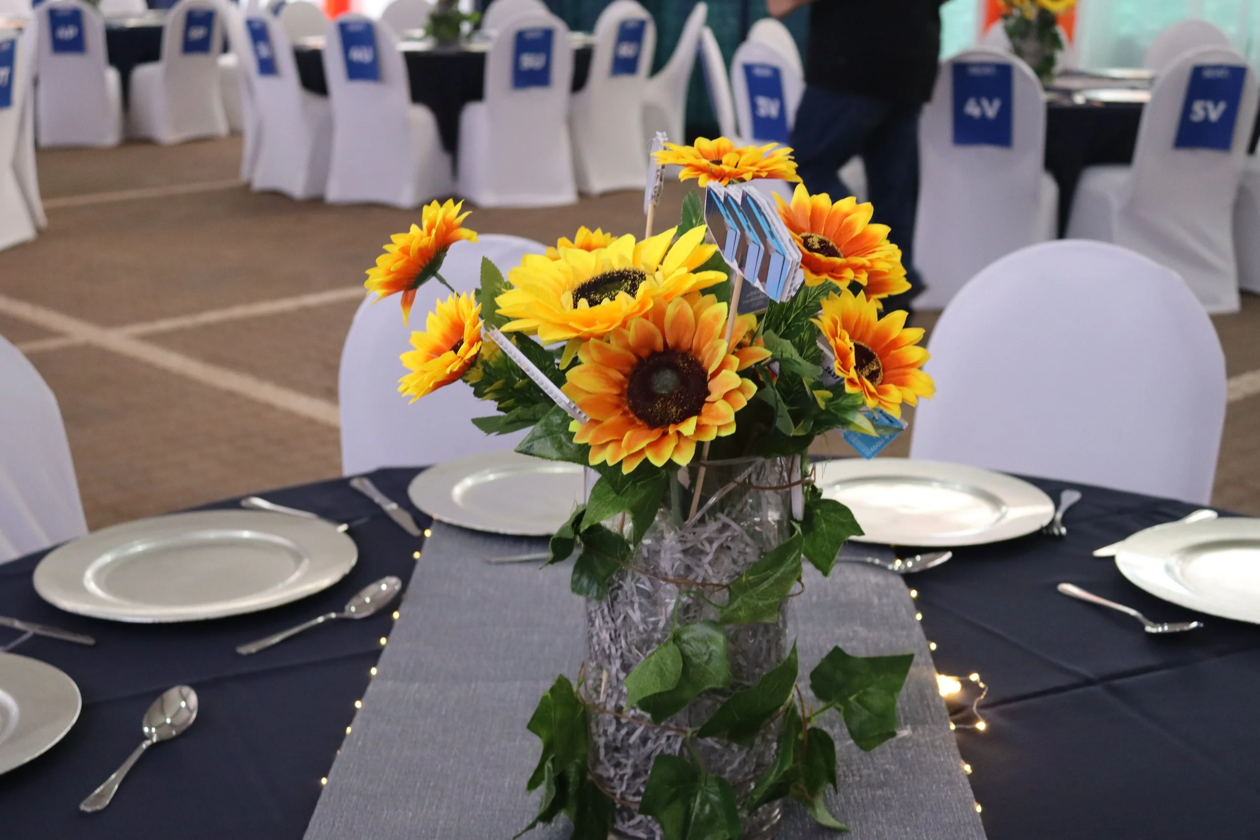 Table centerpiece with bright yellow sunflower flowers, green leaves, and small flags on a dining table with white plates and silverware, set in a decorated banquet hall.