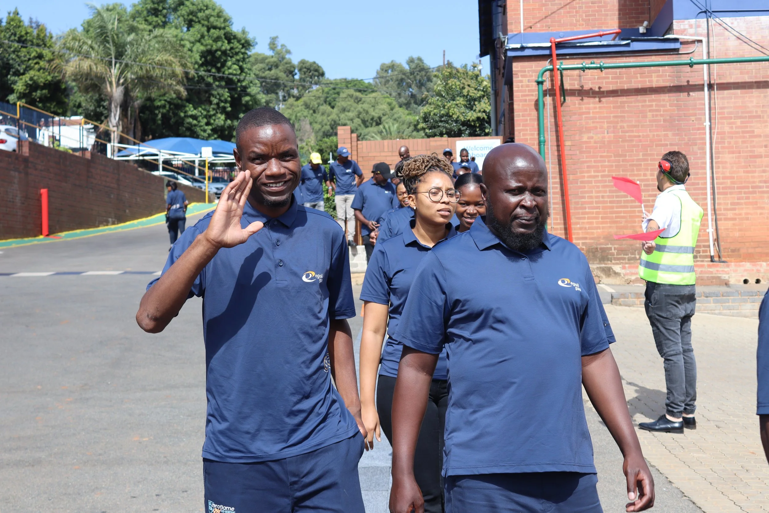 Group of people walking outdoors, dressed in blue shirts, with greenery and brick buildings in the background. One person is waving.