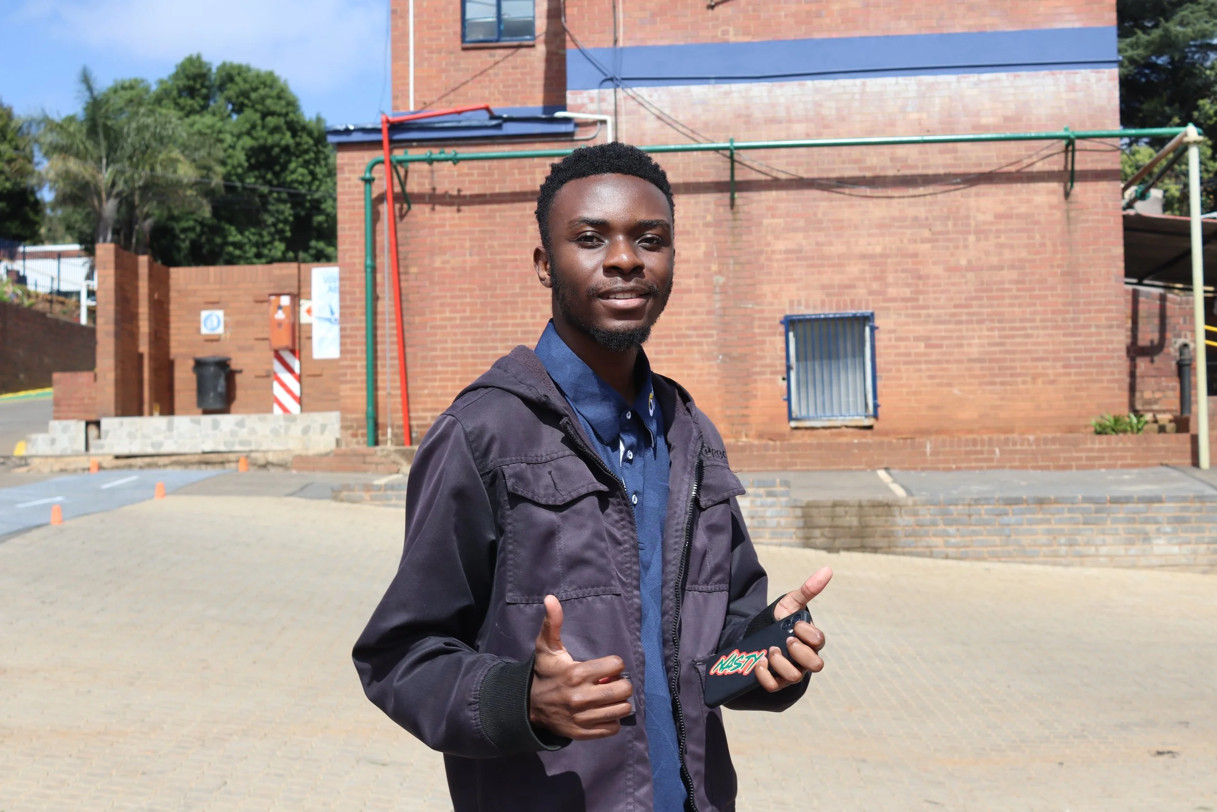 A young man with dark skin and short curly hair standing outdoors in front of a brick building, giving a thumbs-up with both hands, holding a phone in his right hand, and smiling.