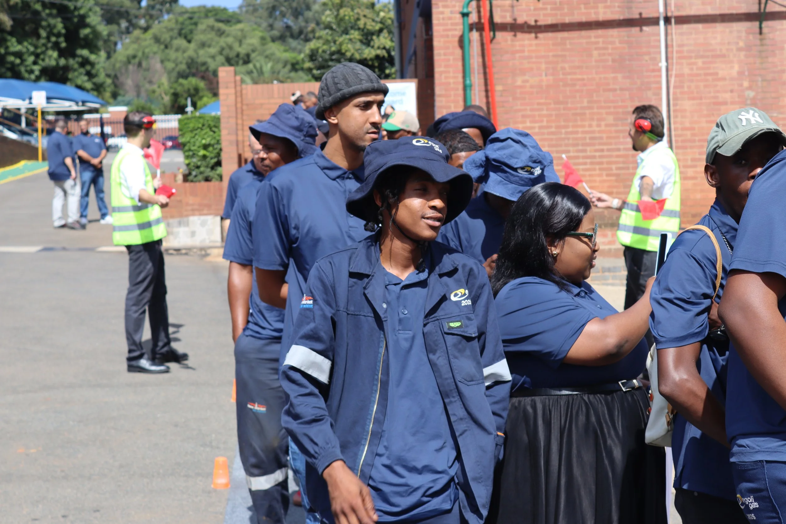 Group of diverse people wearing blue uniforms standing in line outdoors during daytime, with some wearing hats, and a few in the background wearing ear protection and high-visibility vests, near a red brick building.