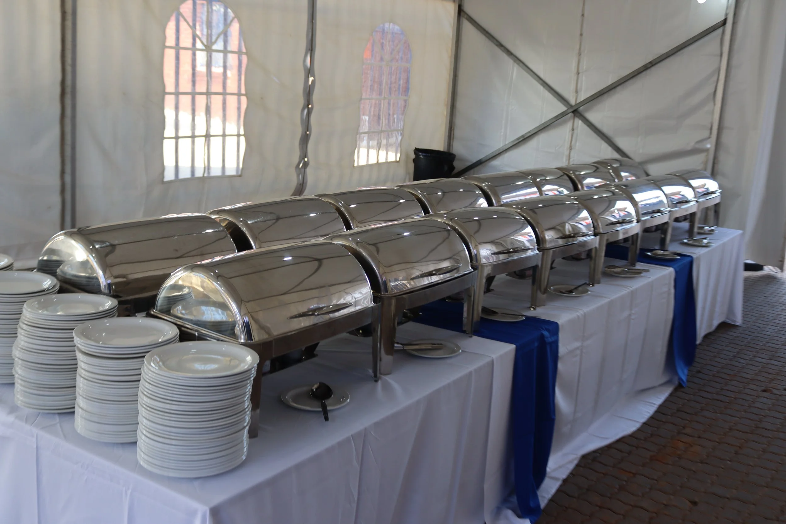 A buffet table with silver chafing dishes and stacks of white plates under a white tent with windows.