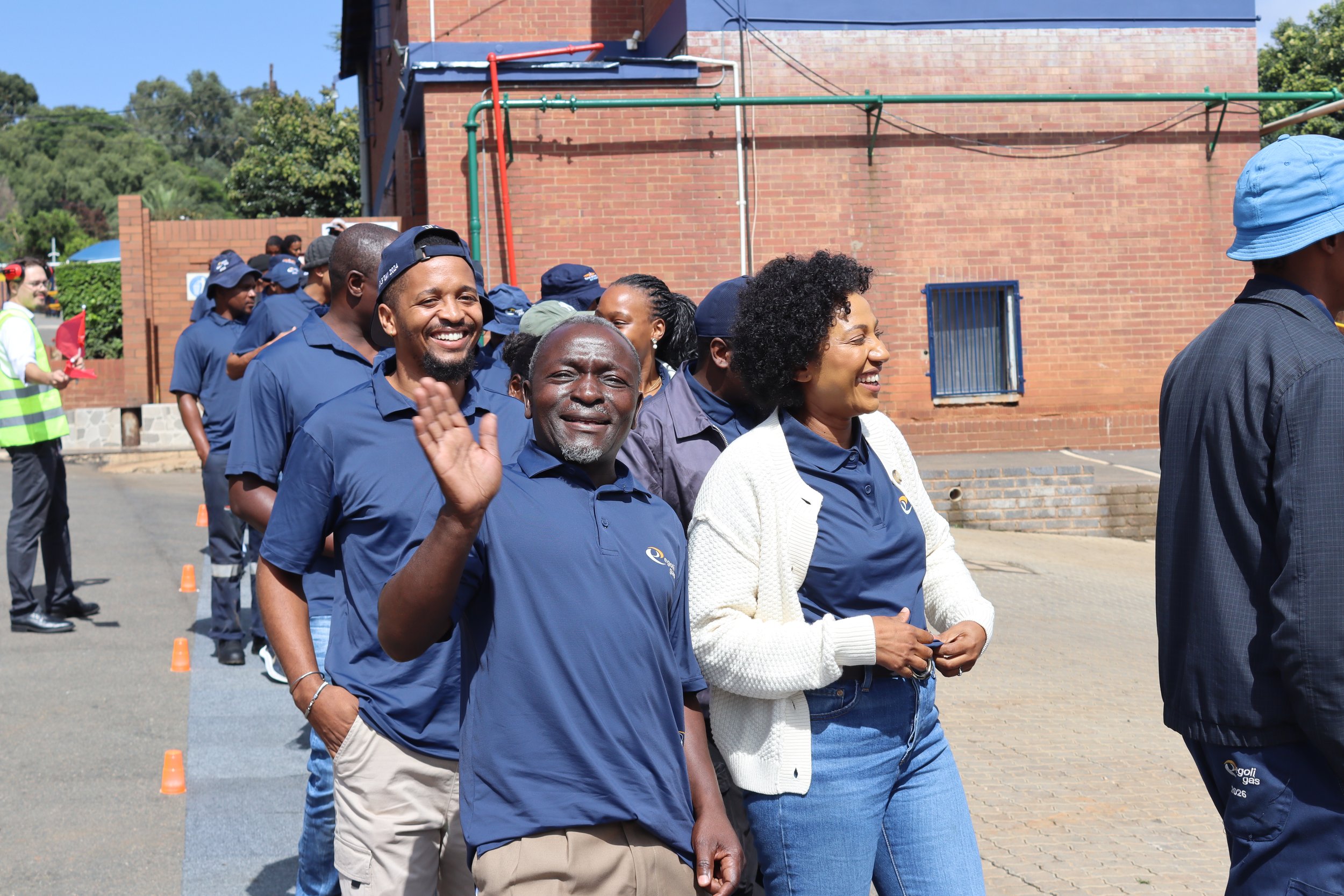 A group of people standing outdoors in a line, some smiling and waving, wearing dark blue shirts, with a brick building and trees in the background.