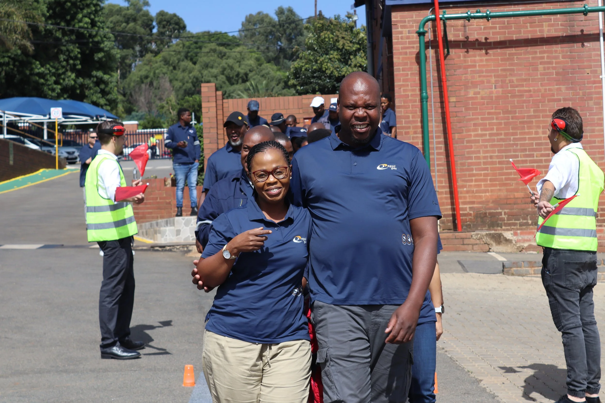 A group of people outdoors, with the focus on a woman with glasses and a man, both smiling and wearing blue shirts, walking together. There are individuals in the background wearing safety vests and holding red flags, in front of a brick building and