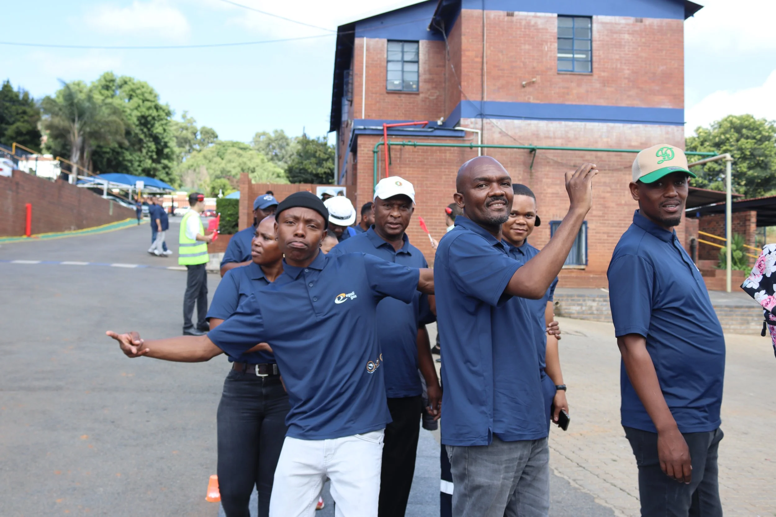 Group of people walking outdoors in front of a brick building, some smiling and waving.