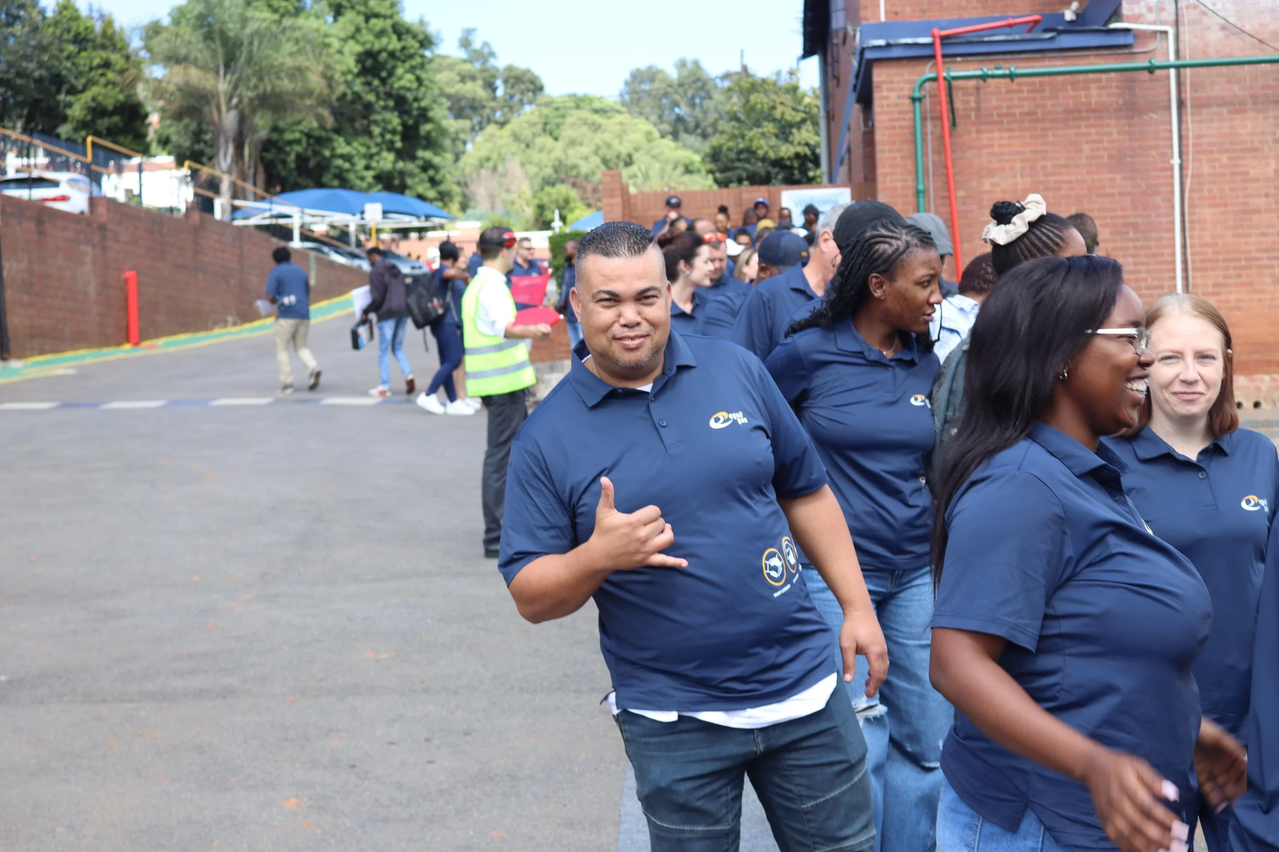 A group of people wearing blue shirts walking outside near a brick building, with trees and parked cars in the background.