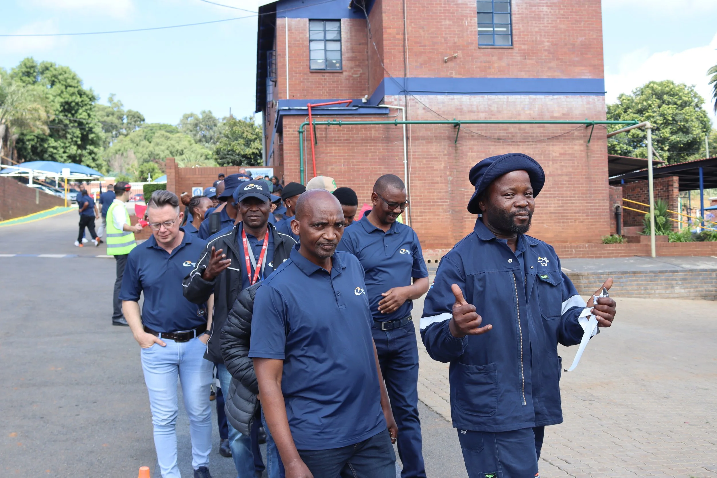 A group of diverse people walking in a line outside near a brick building, some wearing blue shirts and others wearing lanyards, with trees and a blue sky in the background.