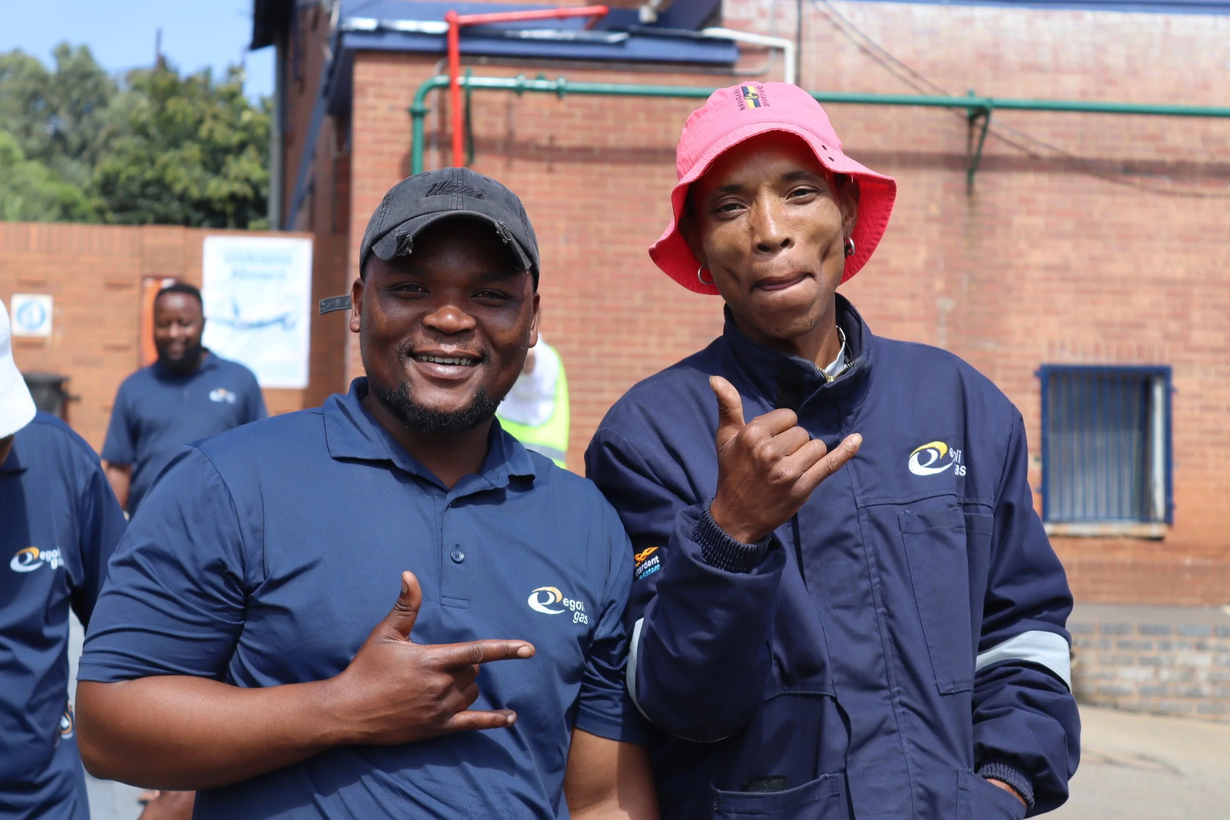 Two men wearing navy blue uniforms and hats, standing outside against a brick building, smiling and pointing with their thumbs.