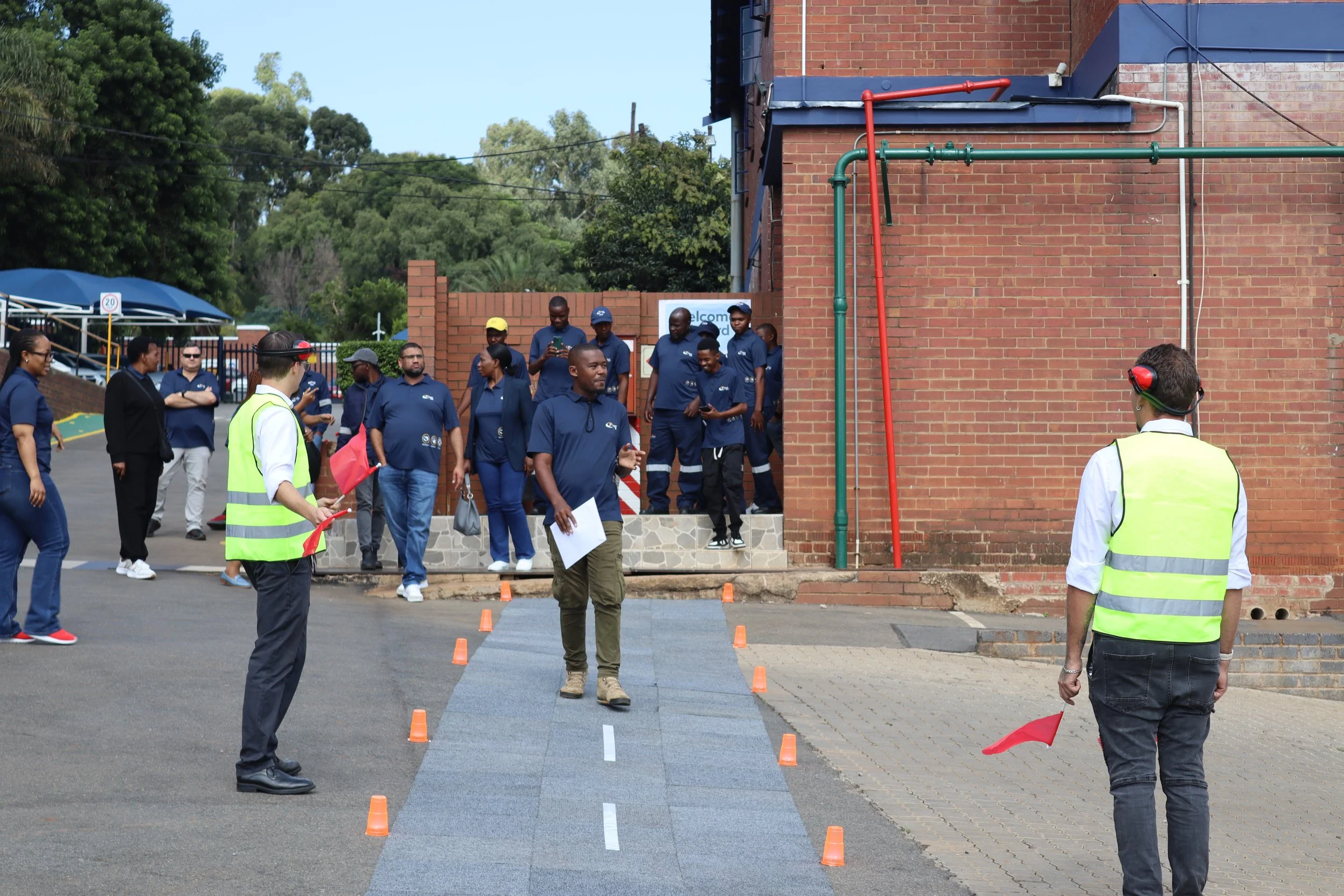 A group of people, some in uniforms, participating in an outdoor event near a brick building. They are standing around a small sectioned-off area marked with orange cones, possibly for a demonstration or training. Some individuals are wearing safety 