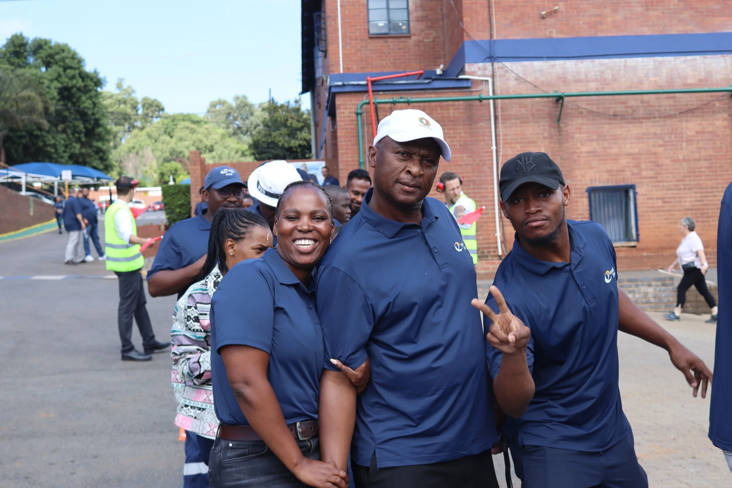 Group of people standing outdoors in front of a brick building, smiling and making gestures, with some wearing caps and reflective vests.