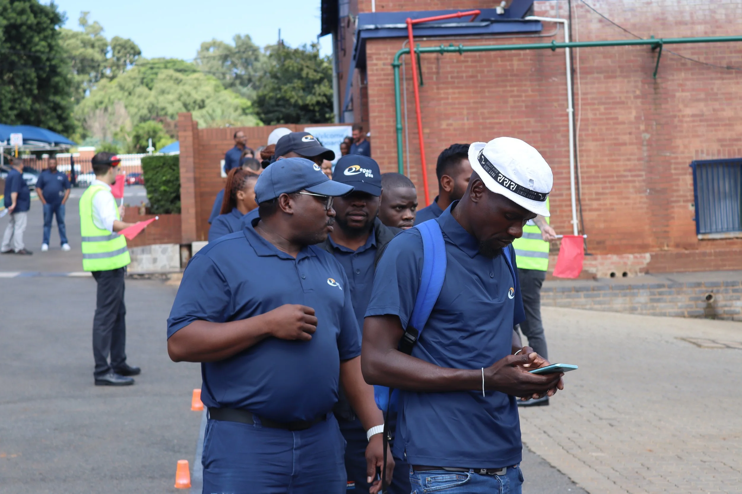A group of people wearing blue shirts and caps, some looking at their phones, gathered outdoors near a brick building with orange and green pipes. Some members are wearing reflective vests, and there are orange cones and a person with a red flag in t