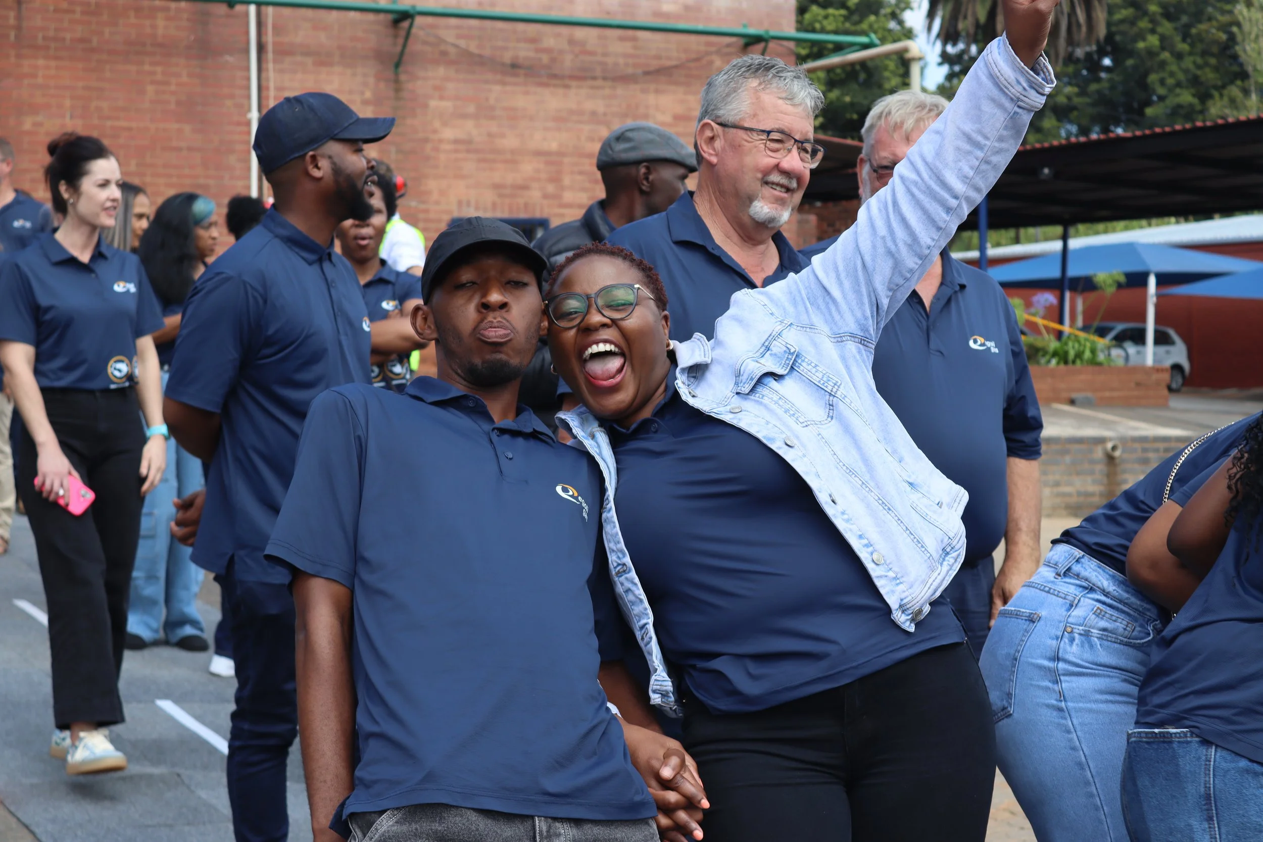A diverse group of people, including a woman and several men of different ethnicities, gathered outdoors during daytime, some smiling and posing for the camera, in front of a brick building.