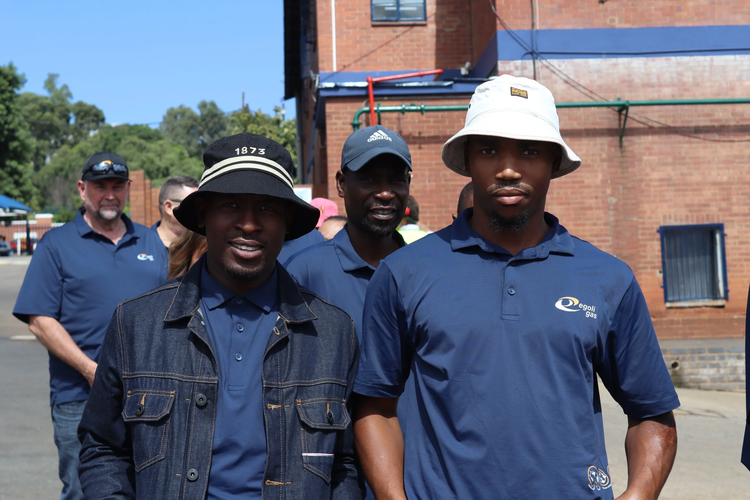 Group of men walking outdoors, some wearing hats, in front of a brick building with utilities and trees in the background.