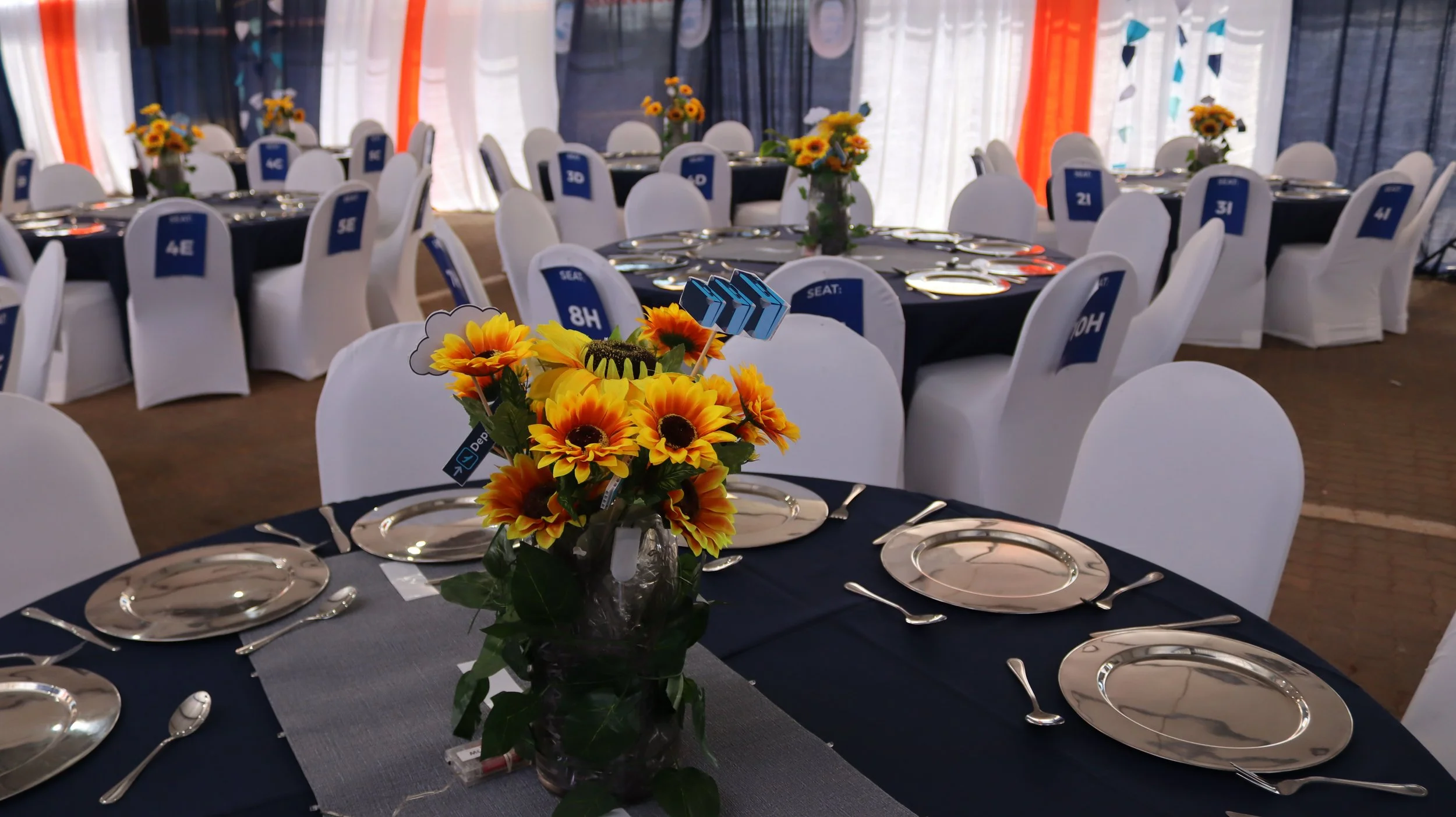 A banquet hall set up for an event with tables decorated with sunflower centerpieces, place settings, and numbered signs for seating, surrounded by white chairs.