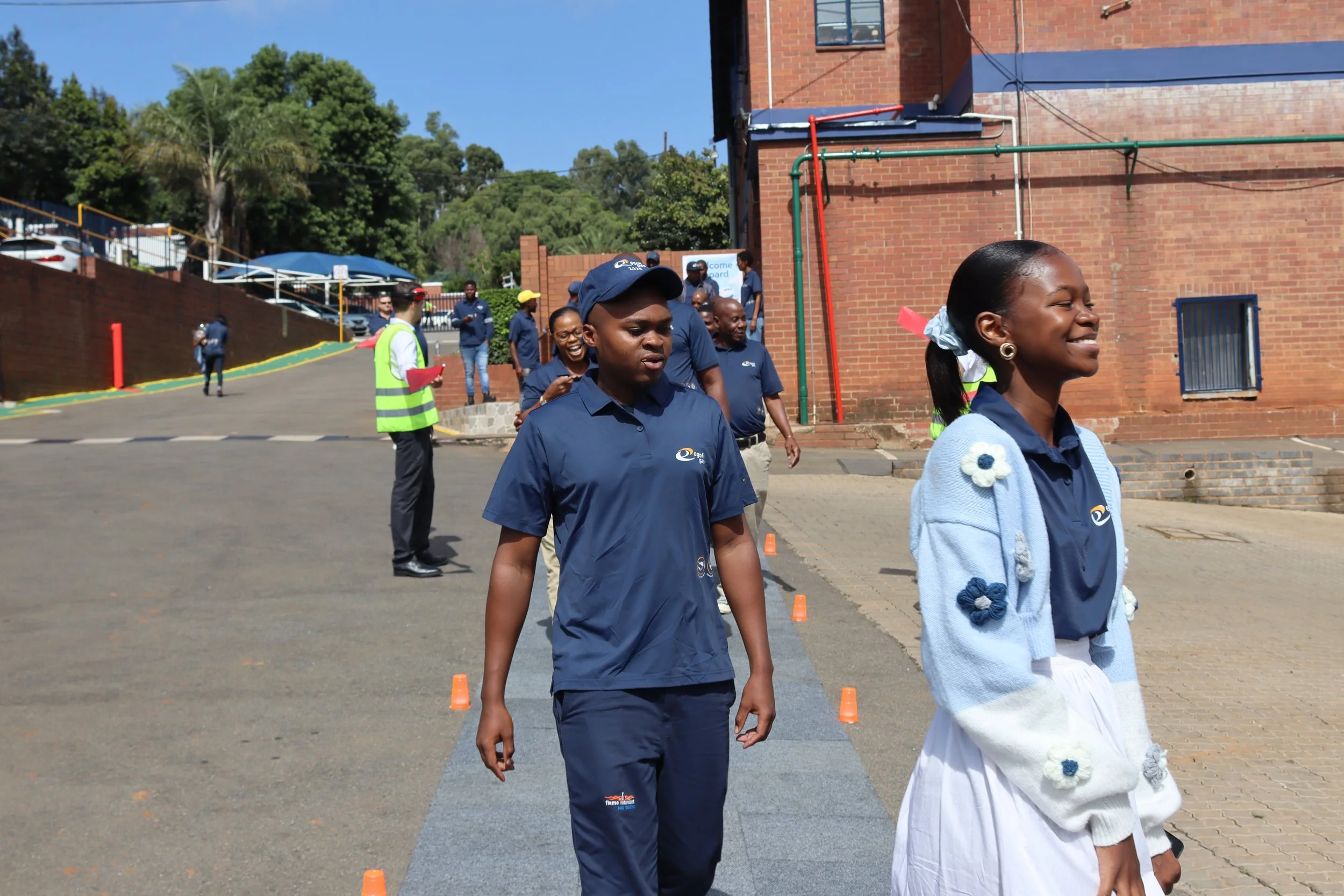 A group of people walking outside near a brick building with colorful pipes, some wearing matching navy blue uniforms, and a woman smiling in the foreground.