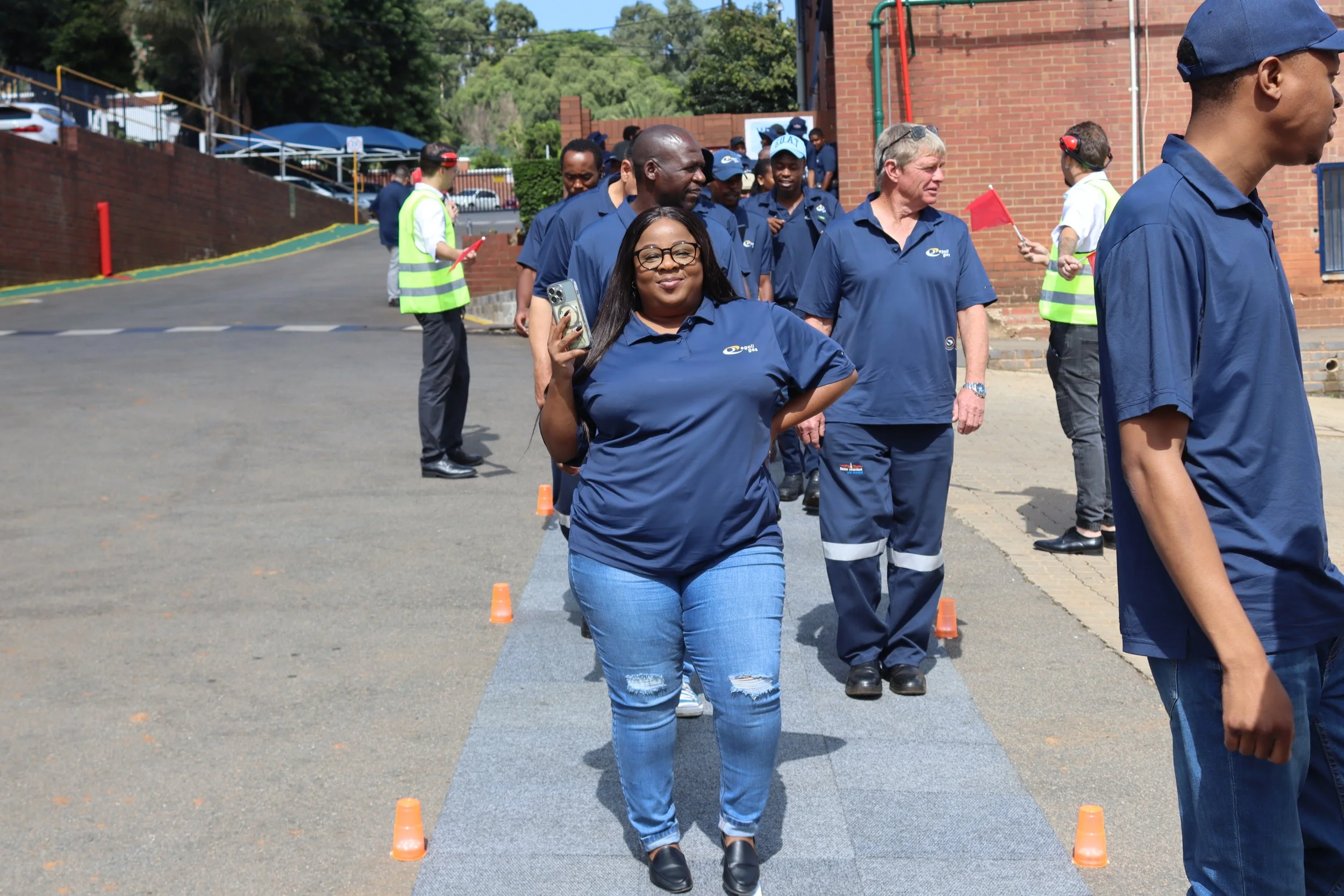 A group of people, predominantly wearing blue uniforms, walking outdoors near a brick building. The woman in the front is smiling, wearing glasses, and holding a phone. Some people in the background are holding red flags, and orange cones are placed 