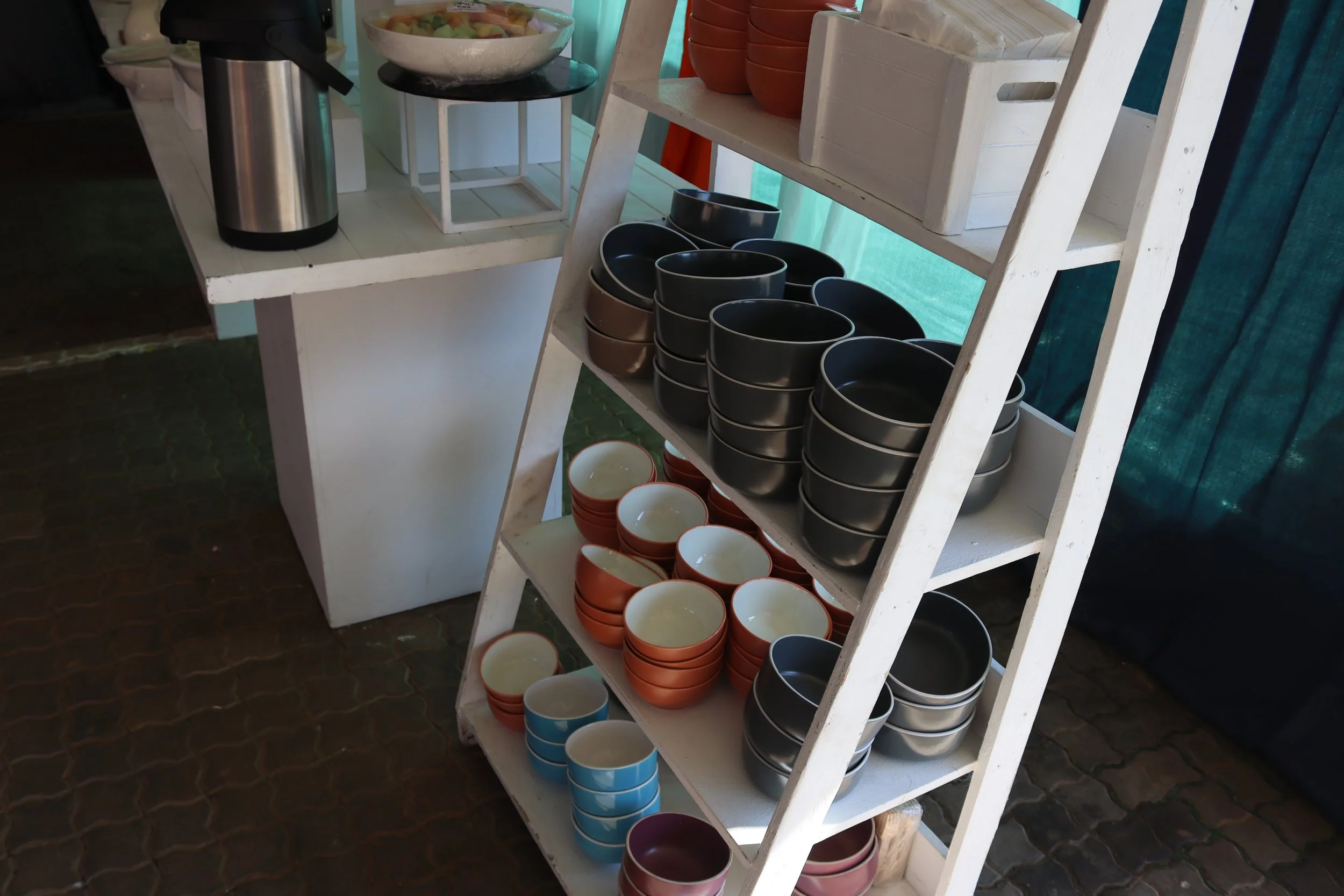 White wooden shelf filled with various bowls and containers, including black, terracotta, and blue bowls, with a background of blue curtain and brick floor.