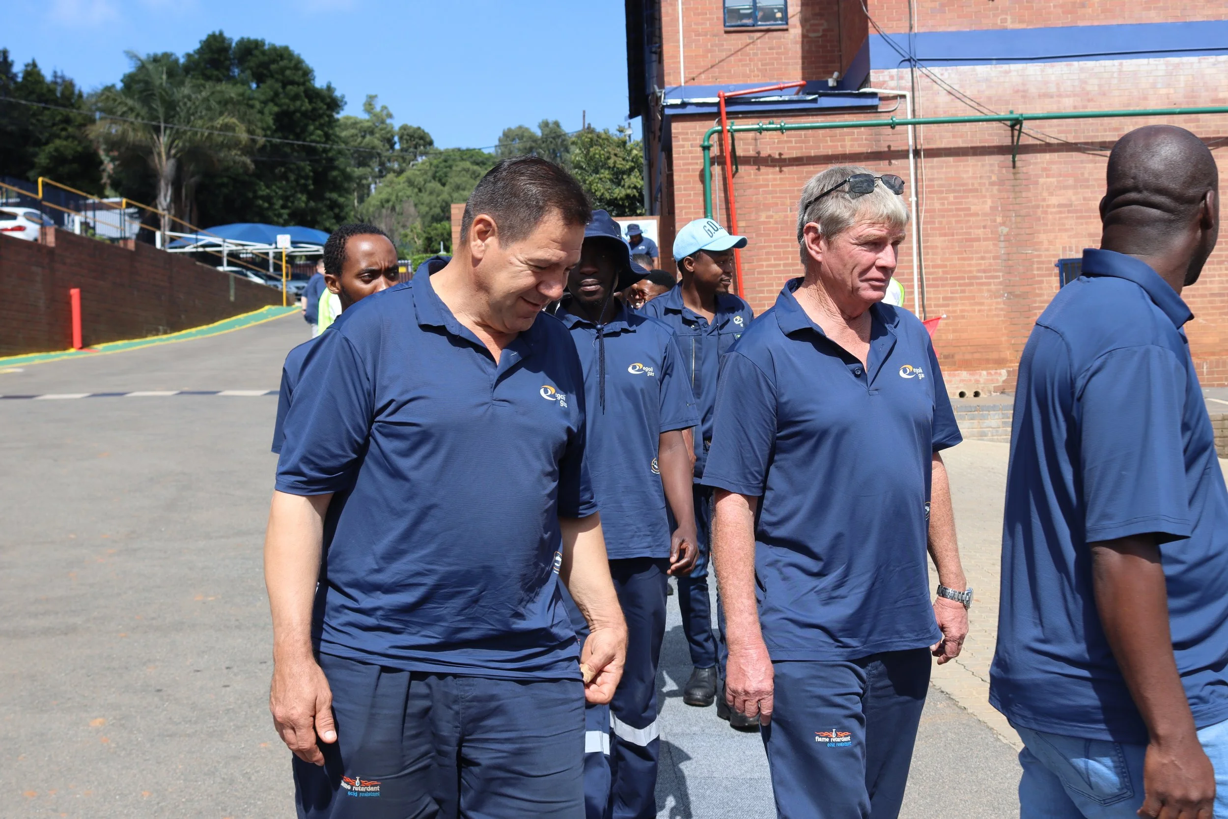 A group of people walking outdoors on a sunny day, wearing matching navy blue polo shirts with logos, near a brick building with trees in the background.