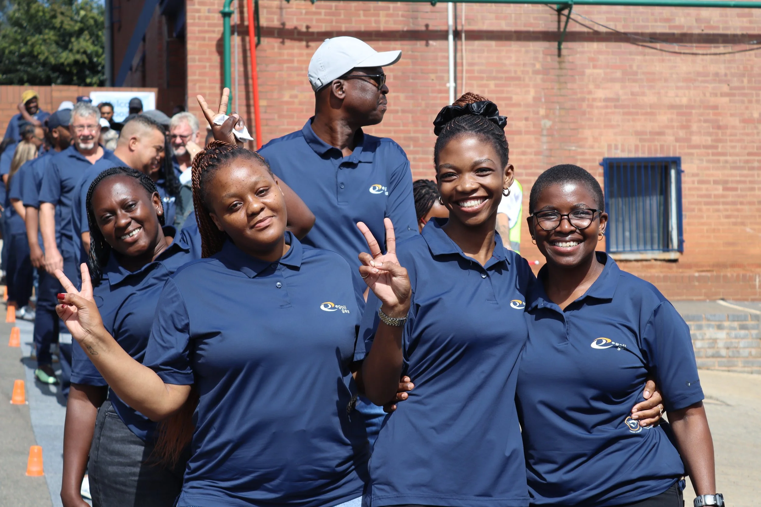Group of smiling women and a man in navy blue shirts, some showing peace signs, during an outdoor event on a sunny day, with a brick building in the background.