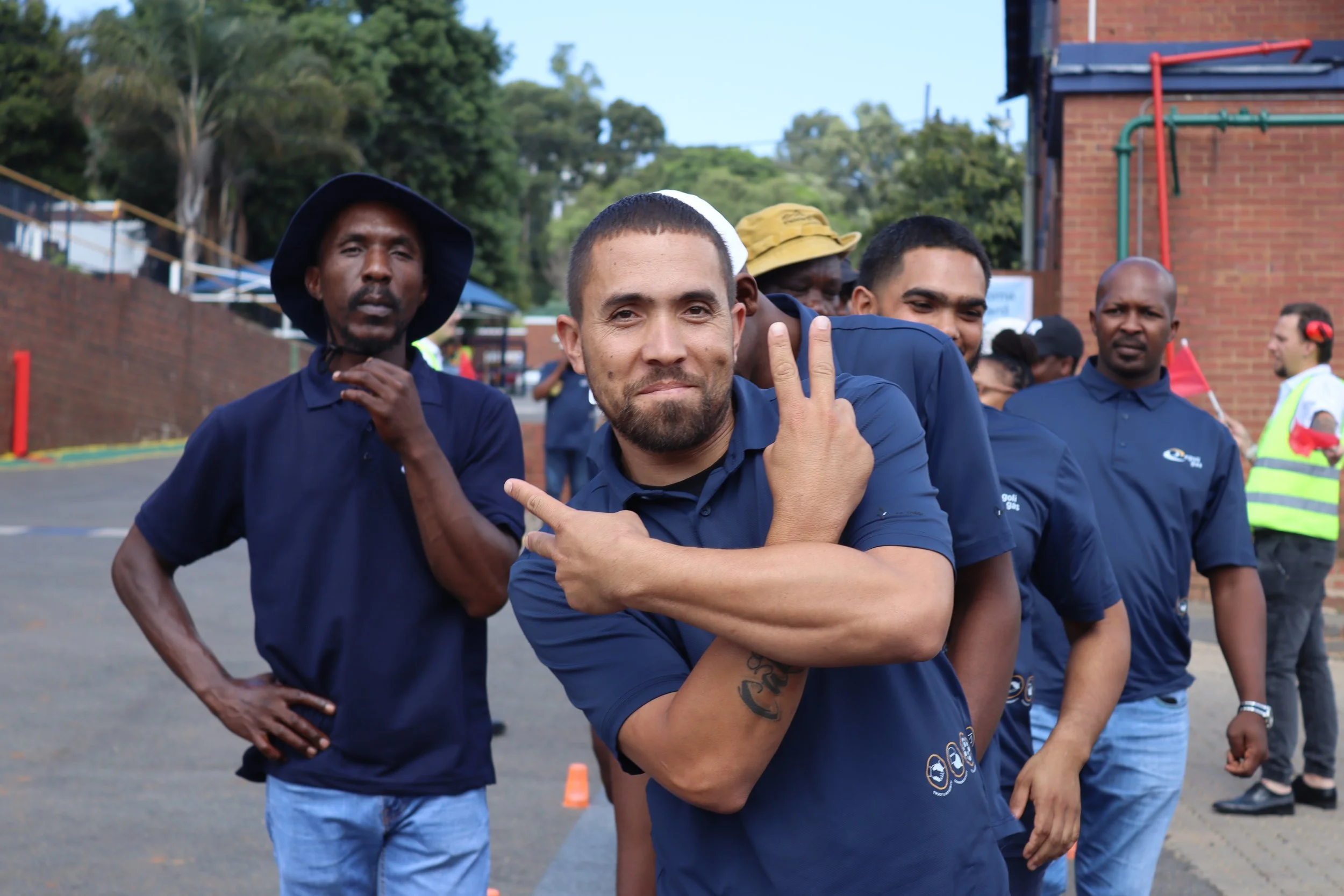 A group of men in blue shirts standing outdoors, some with serious expressions and one smiling while making a peace sign gesture. They appear to be participating in an event or gathering near a brick building with trees in the background.