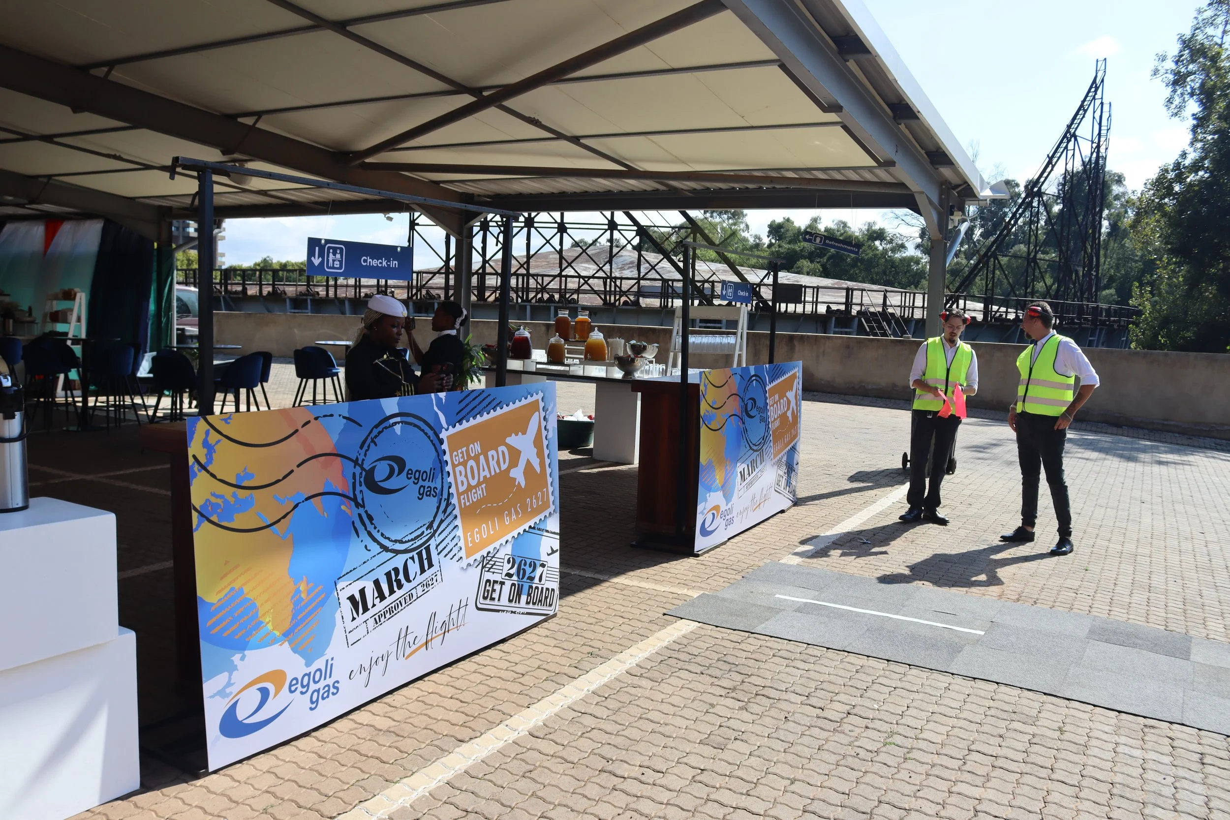 Airport check-in area with two security personnel in reflective vests talking, and a woman at a check-in counter. There are signs indicating check-in and refreshment areas, and promotional banners about Egoli Gas and an event called March 26, 2021.