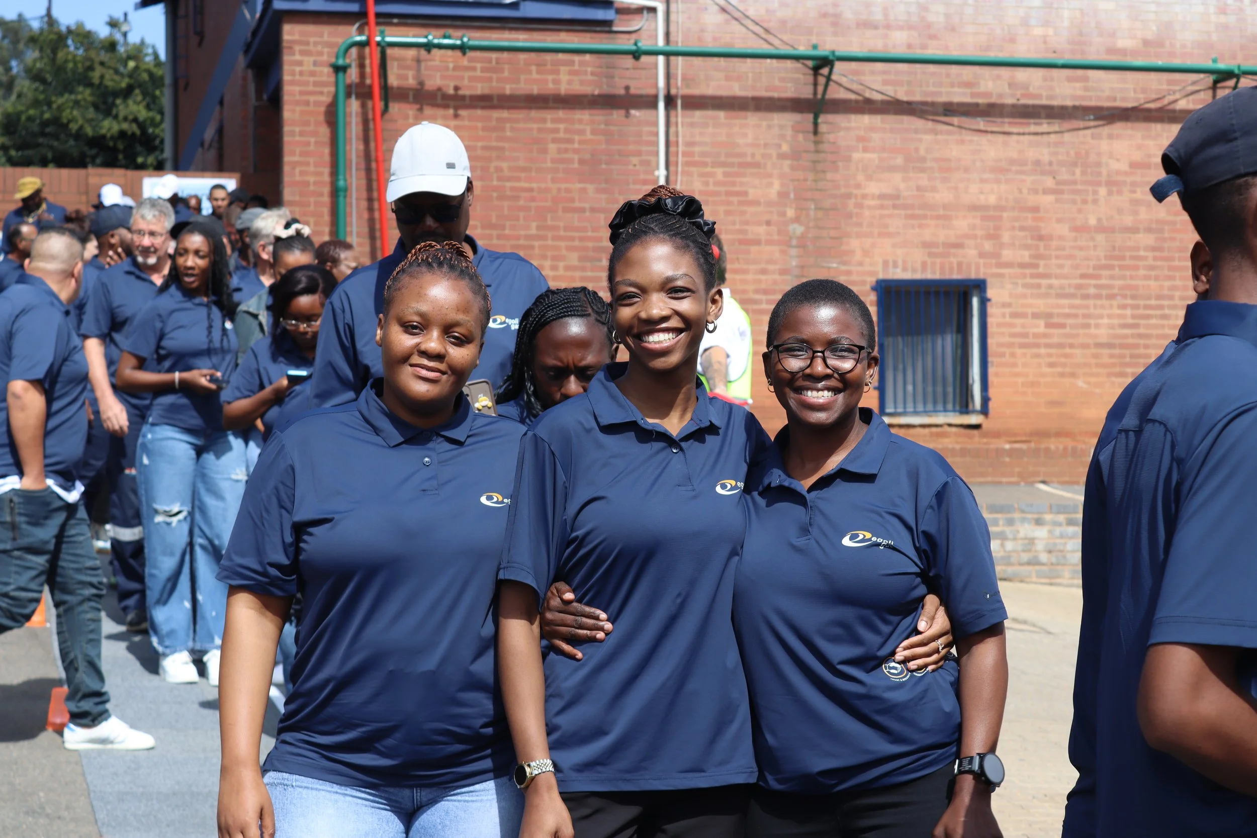 A group of diverse people wearing matching navy blue shirts outdoors, smiling, with some individuals in line in the background against a brick building.