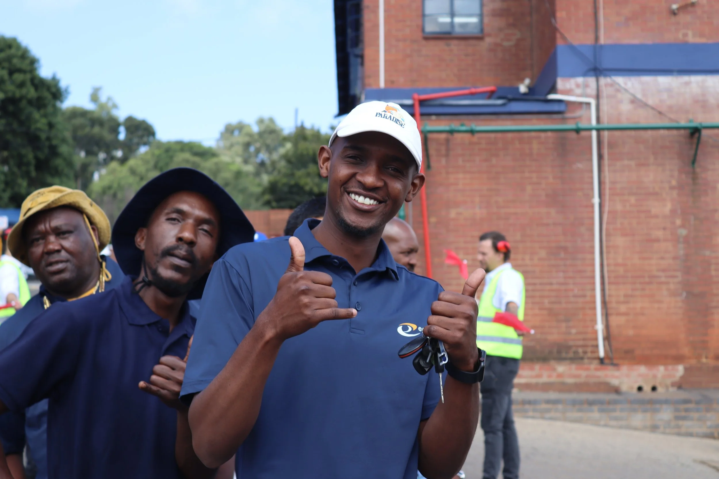 A smiling man wearing a white cap and blue shirt giving a thumbs-up, standing outdoors with a brick building in the background. Other people and greenery are visible behind him.