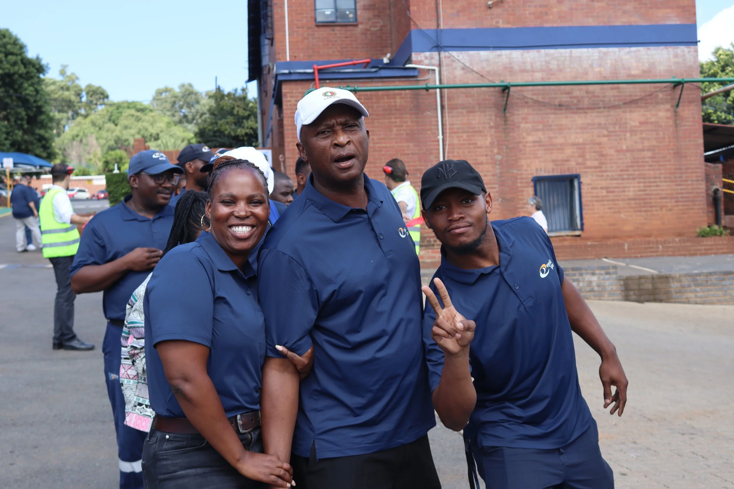 A group of diverse people, wearing blue shirts and hats, poses outdoors in front of a brick building. The group appears happy, with some smiling and making peace signs.