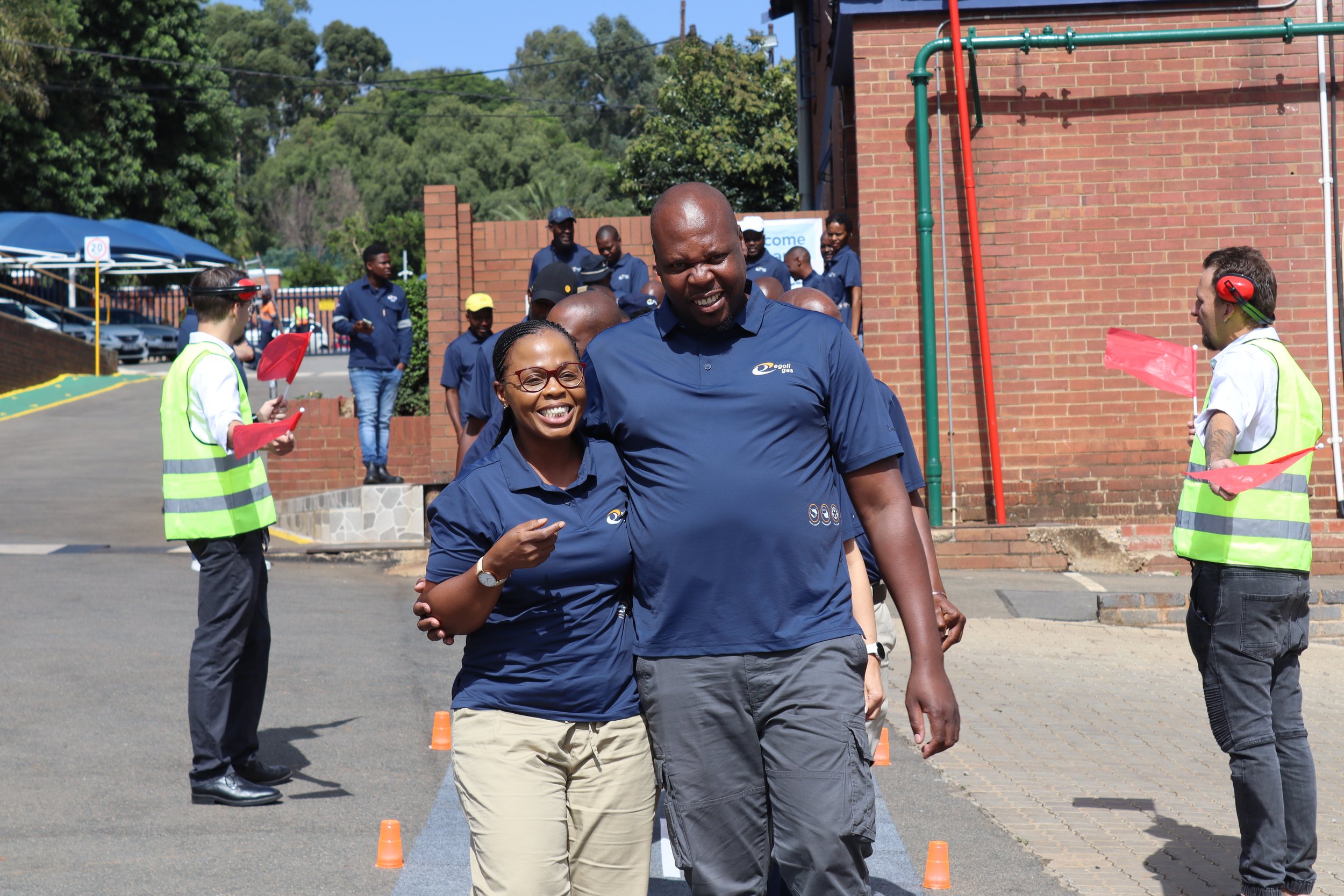 People participating in an outdoor event or race, with some wearing safety vests and others in branded uniforms, near a brick building and green trees on a sunny day.