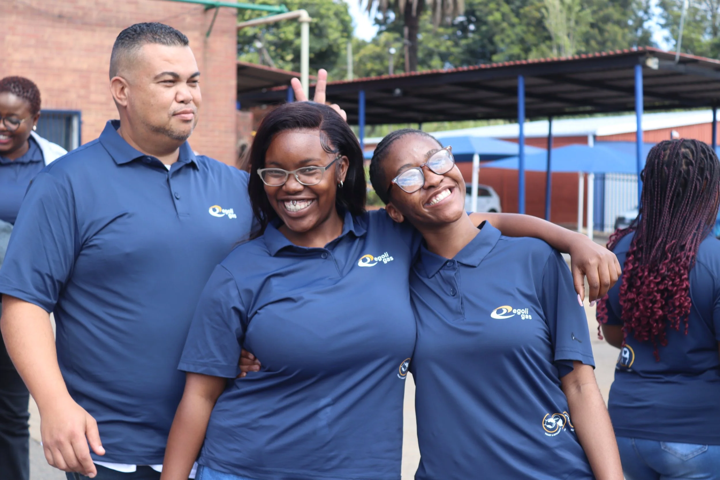 A group of smiling people wearing matching blue shirts, standing outdoors. One person has their arm around another, and two people are making bunny ears behind someone's head. The shirts display a logo that reads 'egoli gas.'