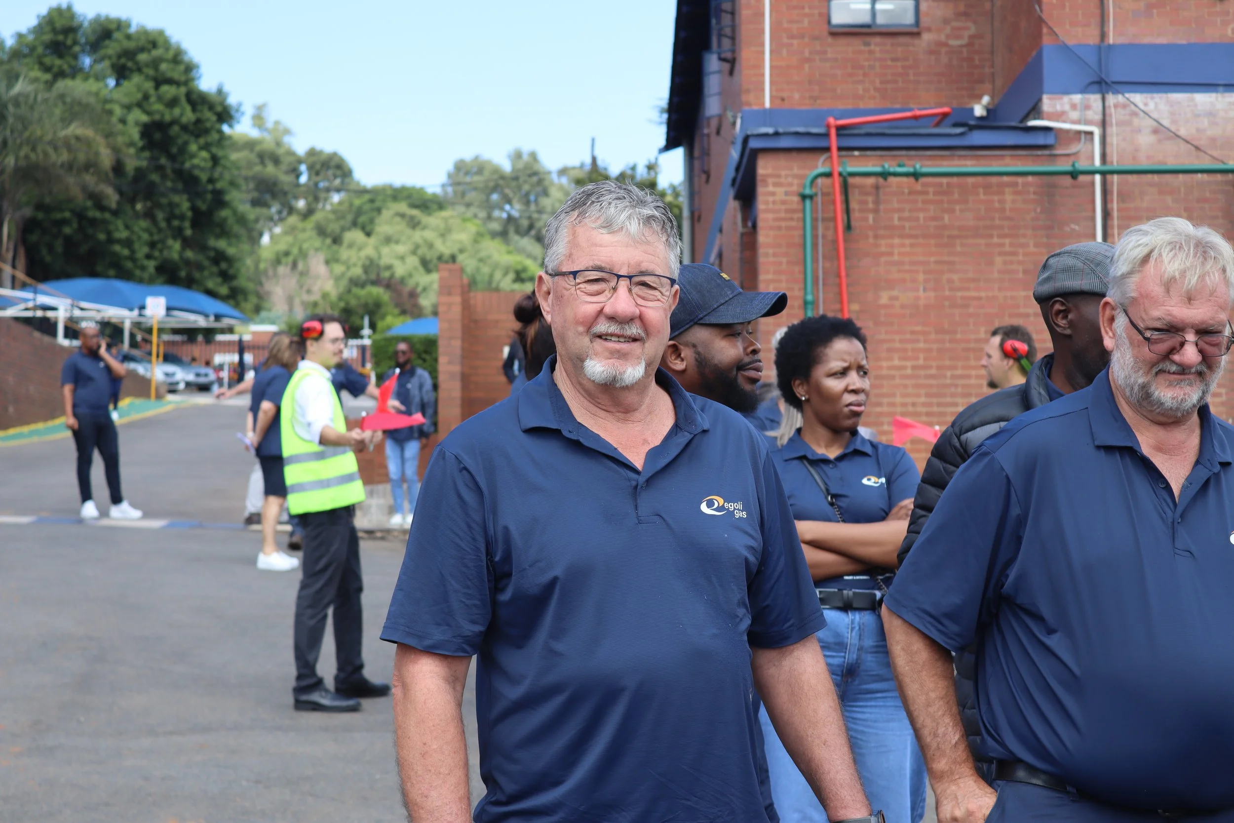 A group of diverse people standing outdoors, some wearing company-branded shirts, in front of a brick building with trees in the background.