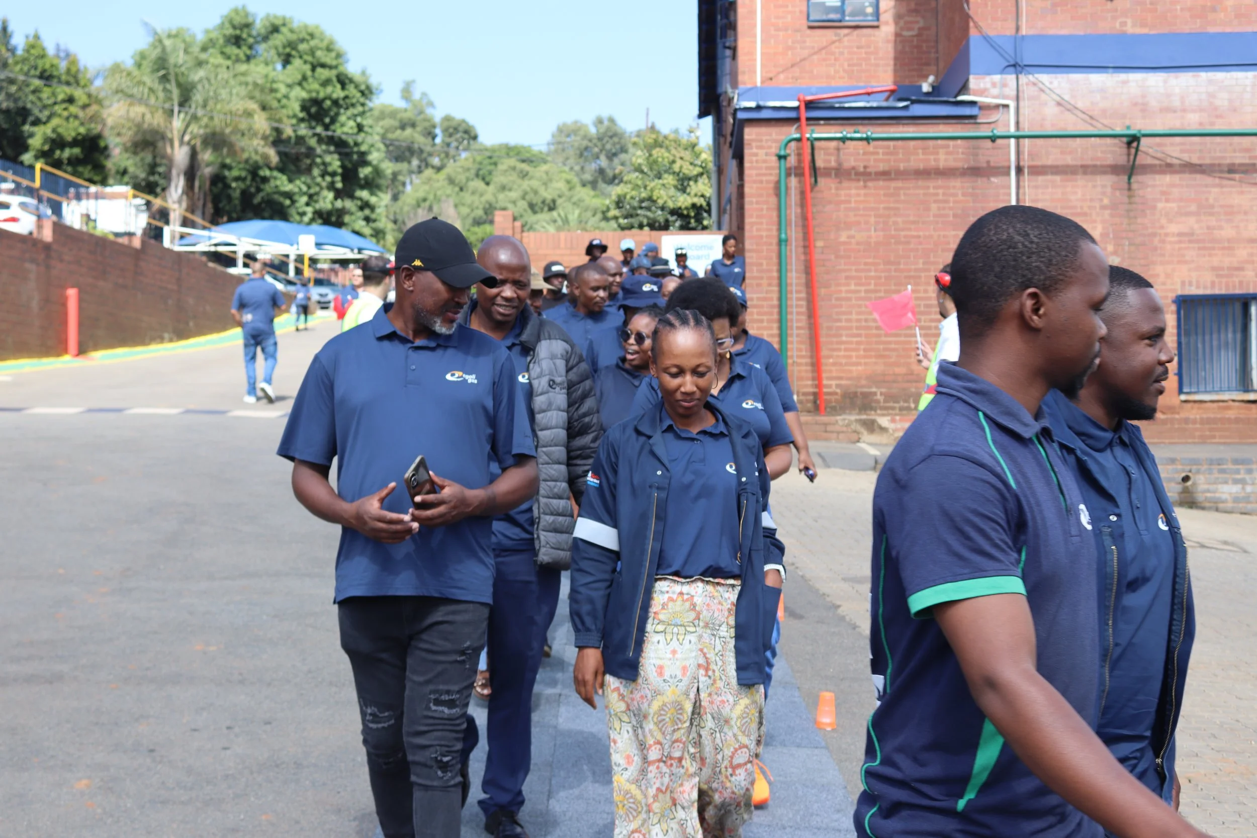 A group of people walking outdoors near a brick building, with some wearing matching blue shirts and one person holding a pink flag.