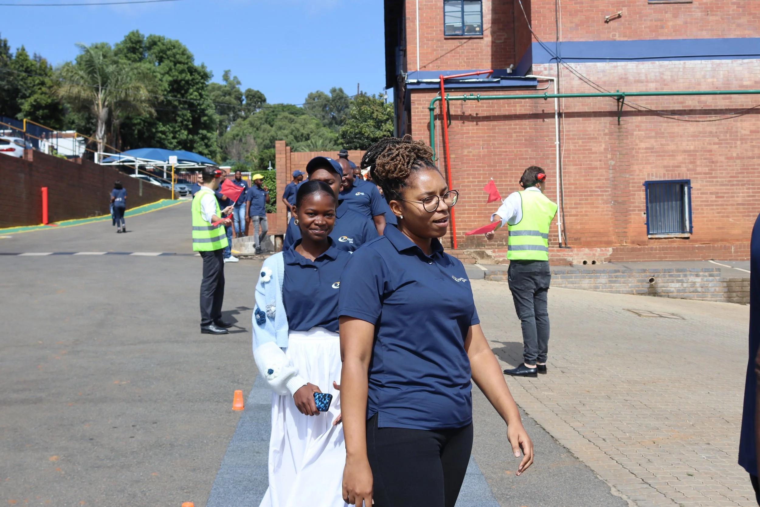 A group of women walking in a queue outdoors beside a brick building, with some holding flags and equipment, with trees and parked cars in the background.