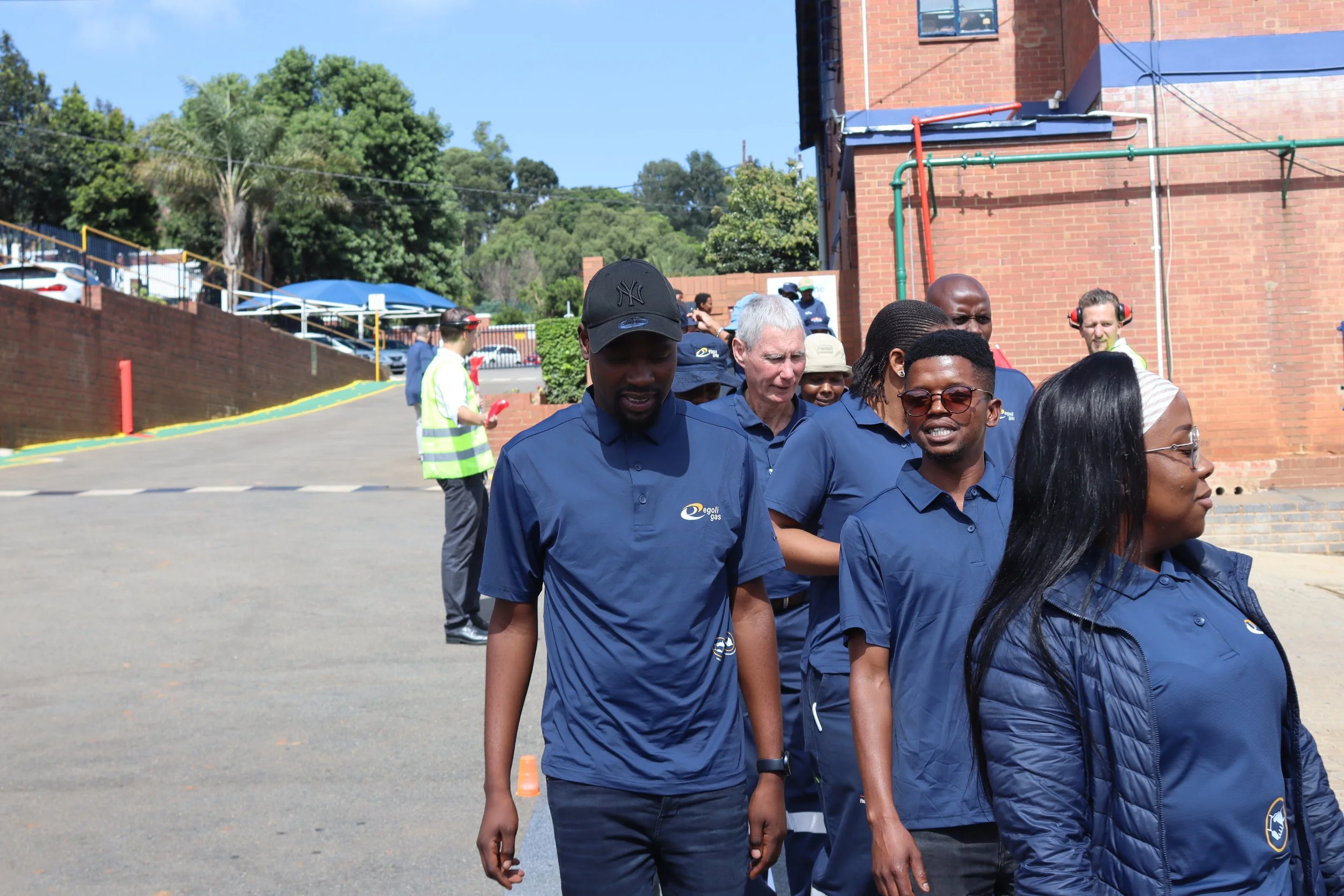 A group of diverse people walking outdoors near brick buildings, some wearing uniforms with logos, under a clear sky.