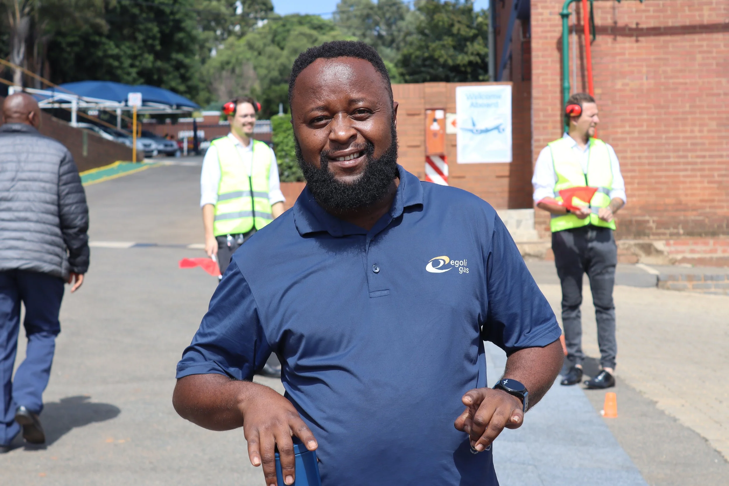 A smiling man with a beard in a blue polo shirt standing outdoors with people in the background. Others are wearing safety vests and headphones, suggesting an event or work setting near a brick building.