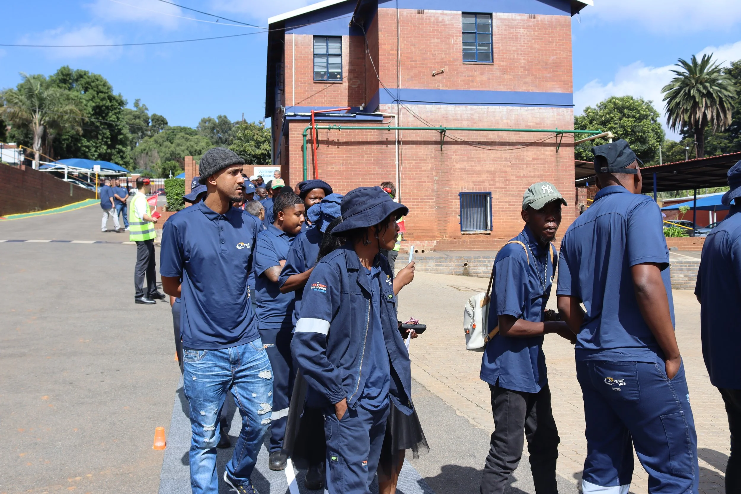 People in line outdoors near a brick building on a sunny day, some wearing blue uniforms and hats, with trees and blue sky in the background.