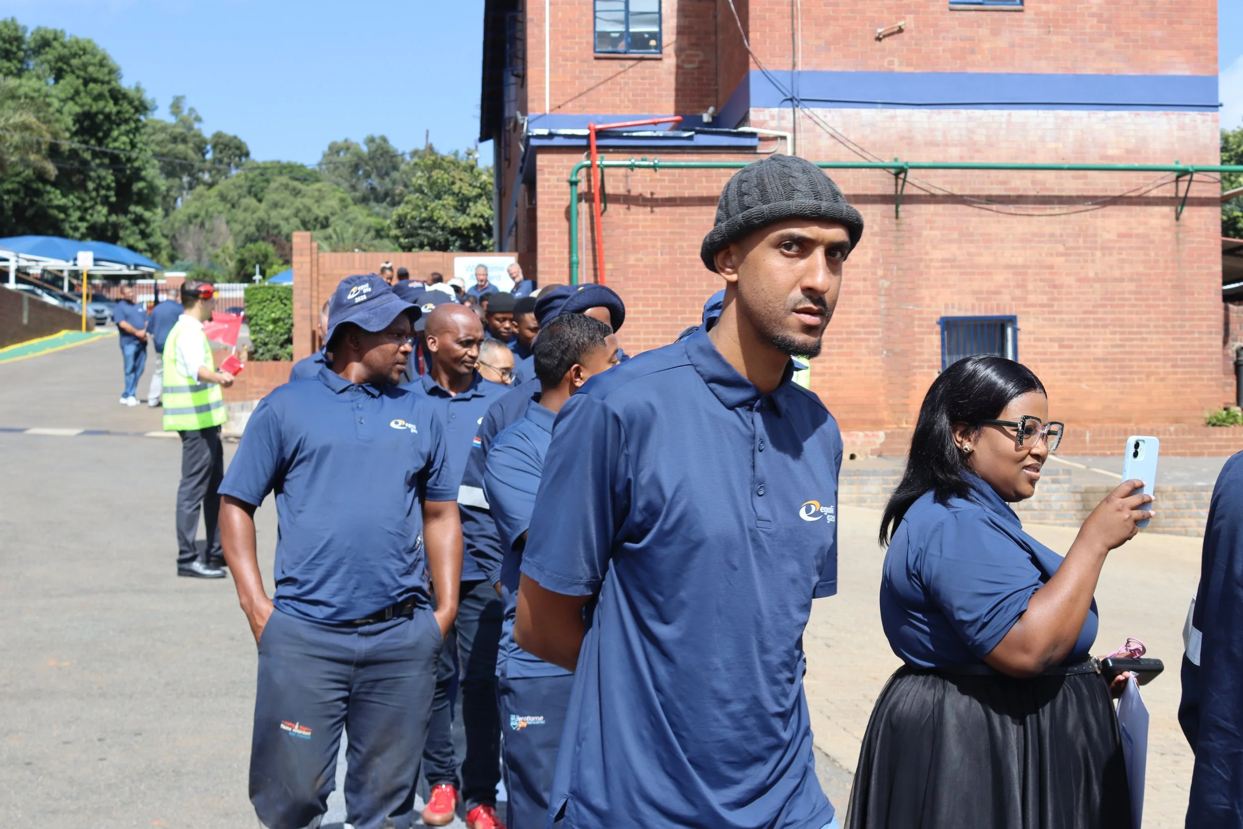 A group of people dressed in blue shirts standing in line outside near a brick building, with some taking photos or looking at their phones, on a sunny day.